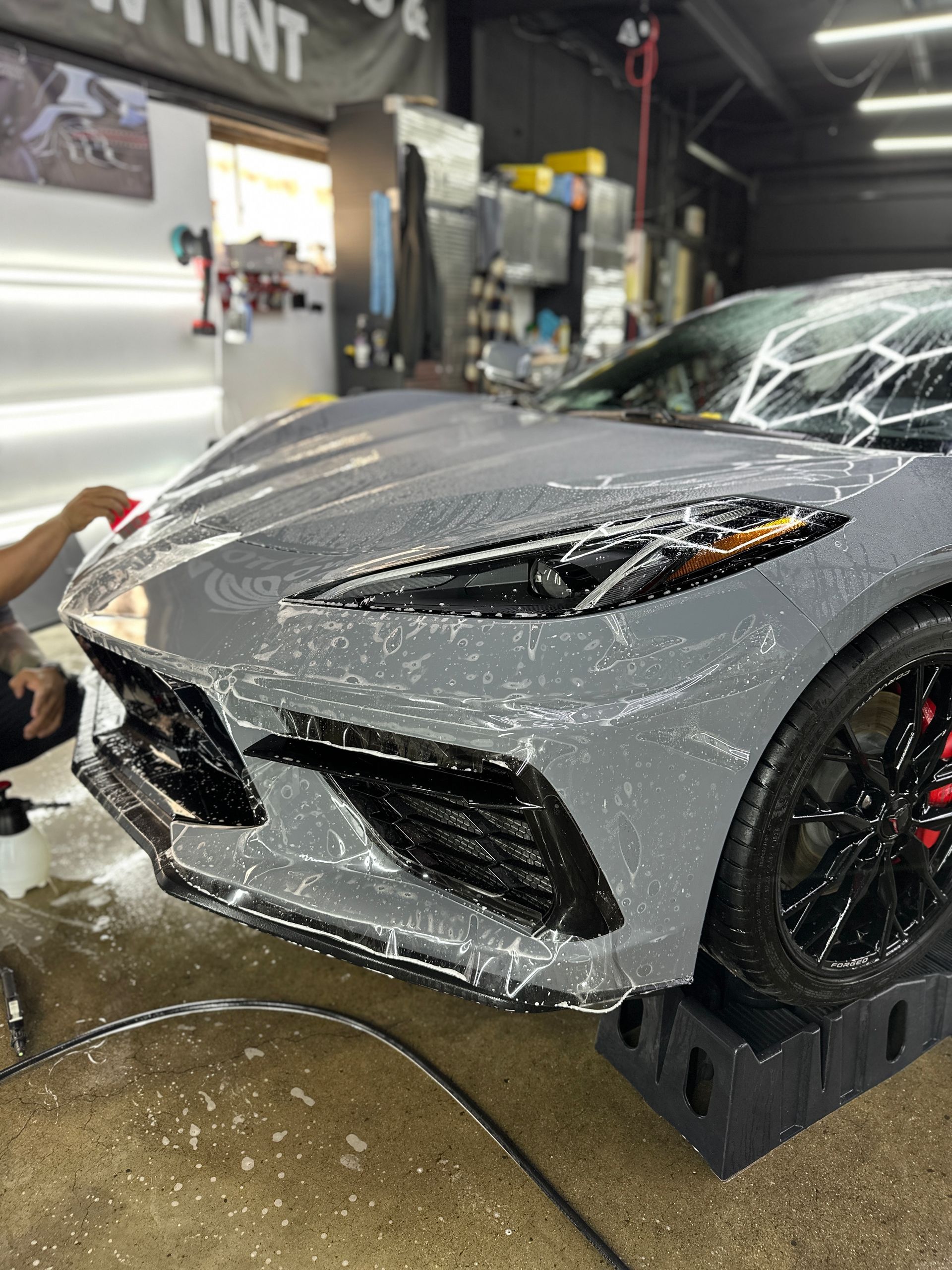 A grey sports car being washed with soap in an auto shop with the front wheel raised on a ramp.