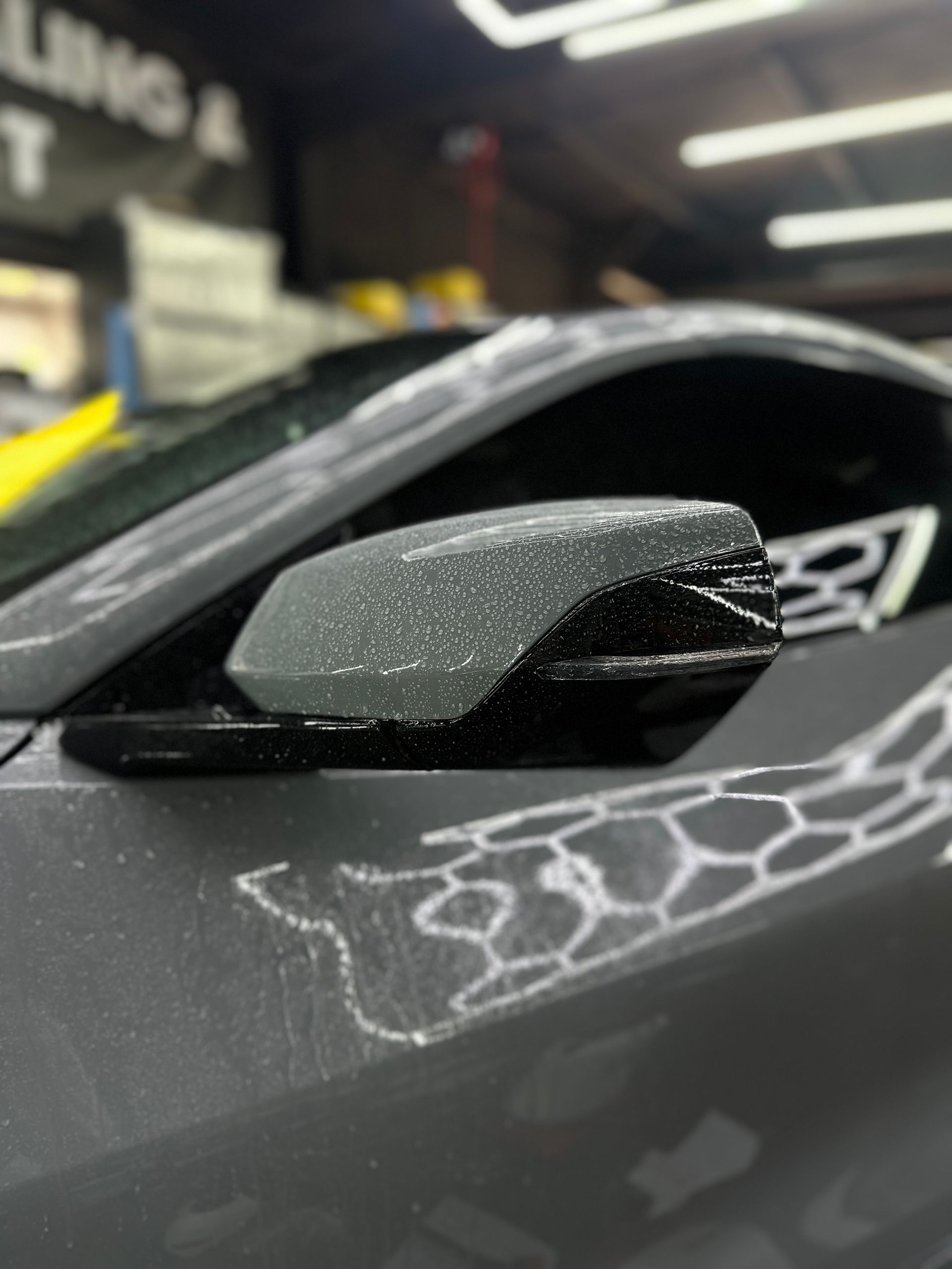 Close-up of a car side mirror with a matte grey finish, black accents, and a white geometric hexagonal pattern.
