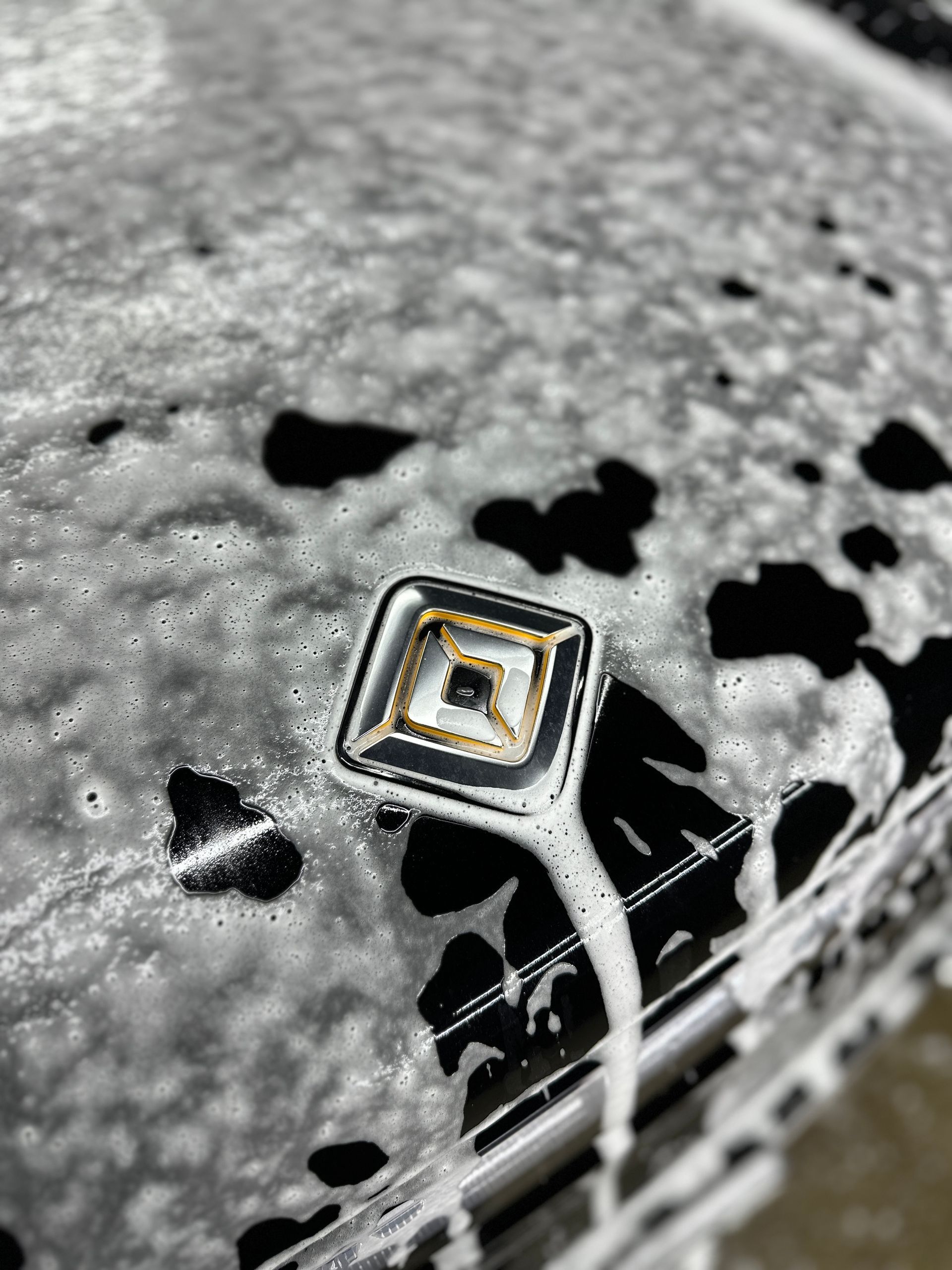 A square, silver-rimmed emblem with a patterned center, partially covered in soapy foam on a car hood.