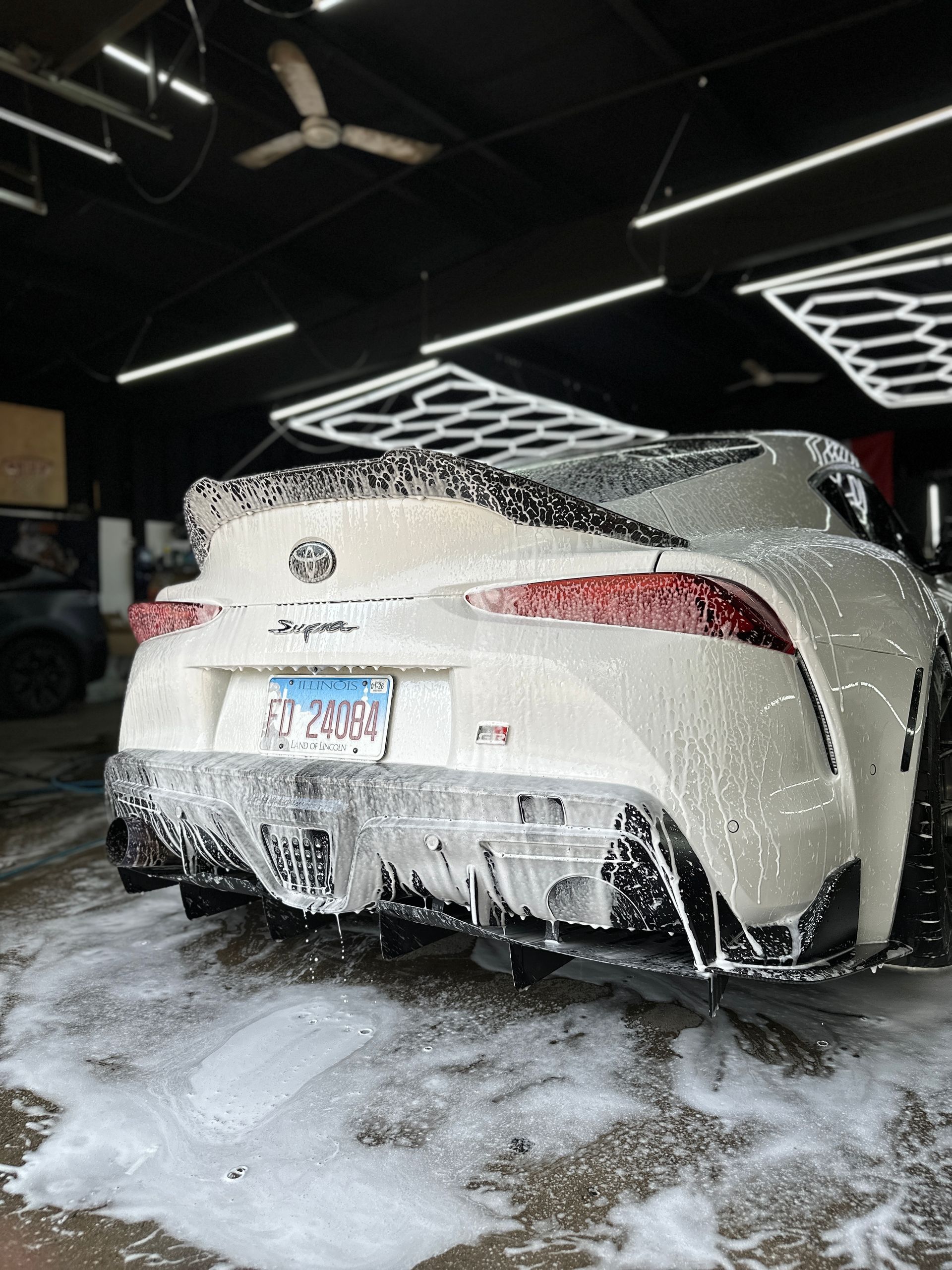 A white Toyota Supra covered in car wash soap, parked in a well-lit, industrial-style garage.