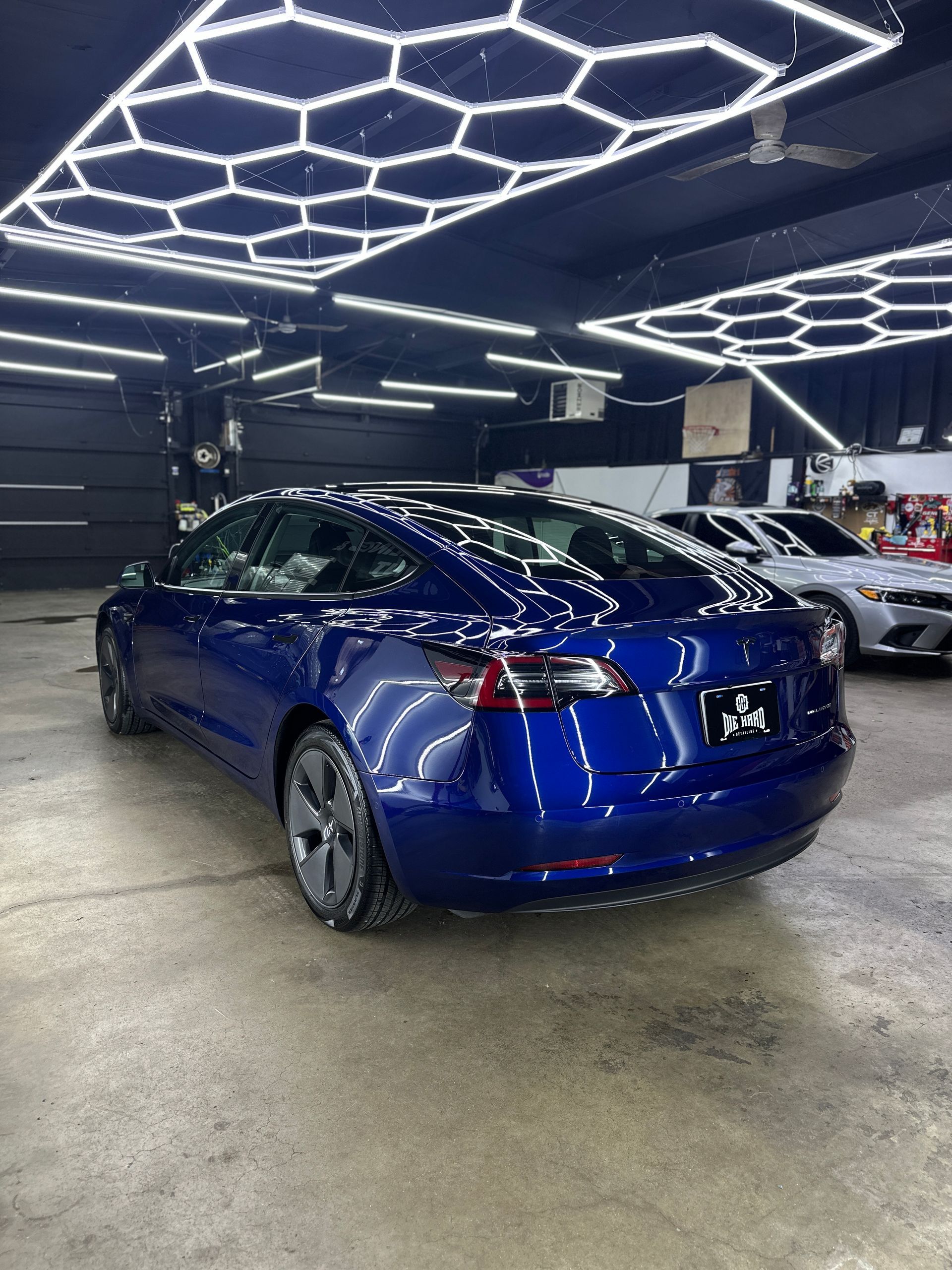 A shiny deep blue Tesla parked in a professional detailing shop with hexagon ceiling lights and an industrial floor.