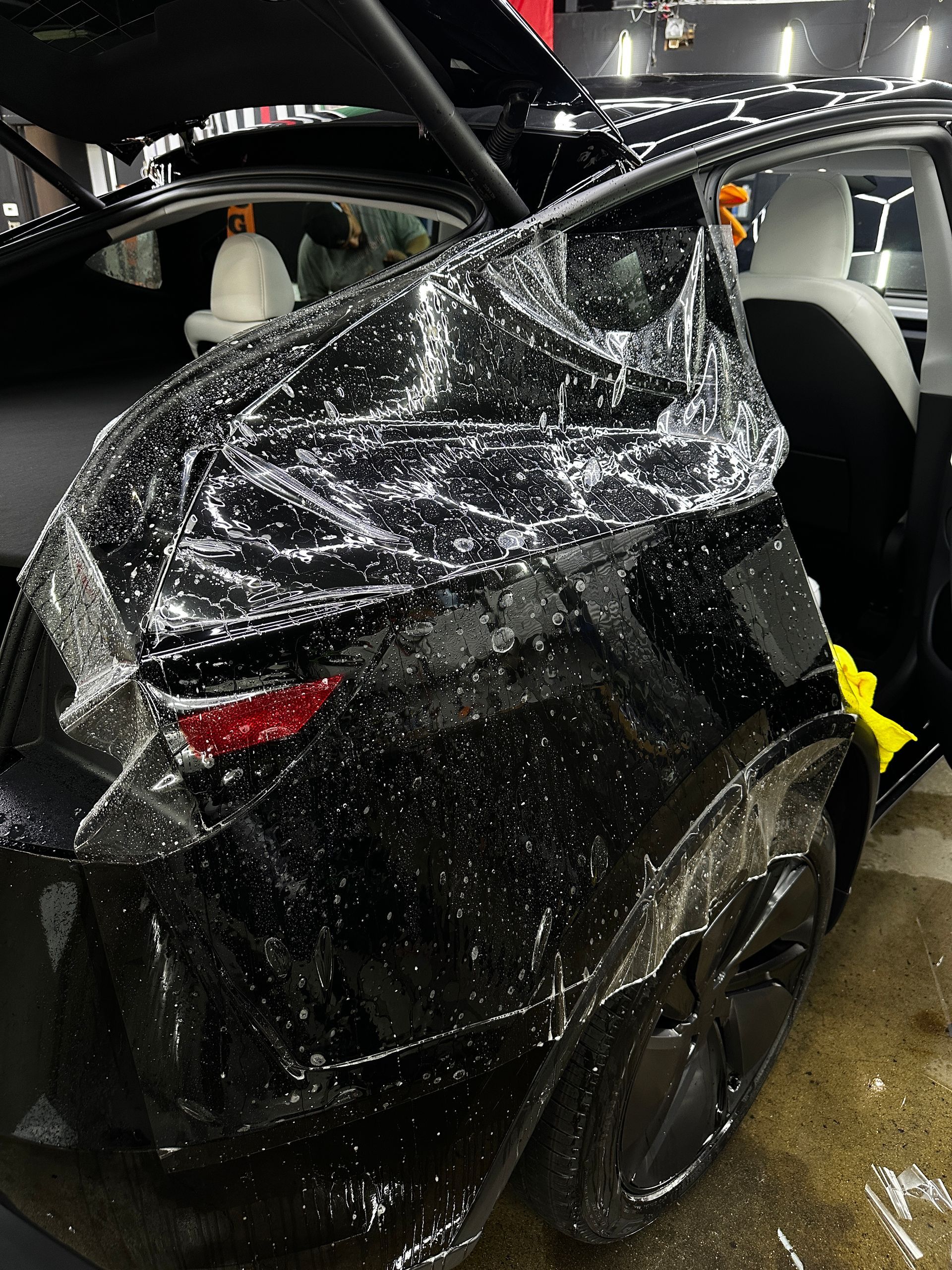 A black car with protective plastic film applied to its rear quarter panel and door in an indoor garage.