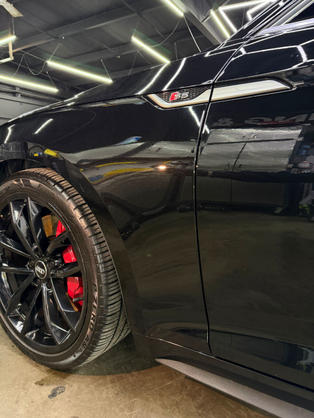 A black Audi S5 with a red brake caliper, shown from the front wheel well in a workshop setting.