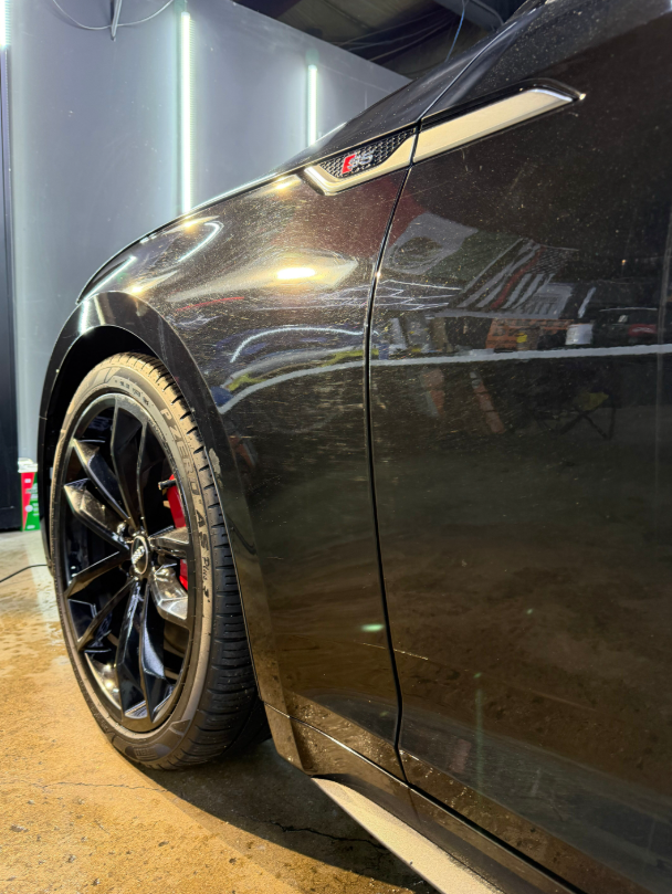 Close-up of a black Audi sports car showing the front wheel, red brake caliper, and side fender trim in a garage.