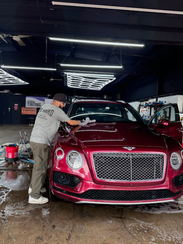 A technician in a grey t-shirt cleans the windshield of a metallic dark red Bentley inside a well-lit auto shop.