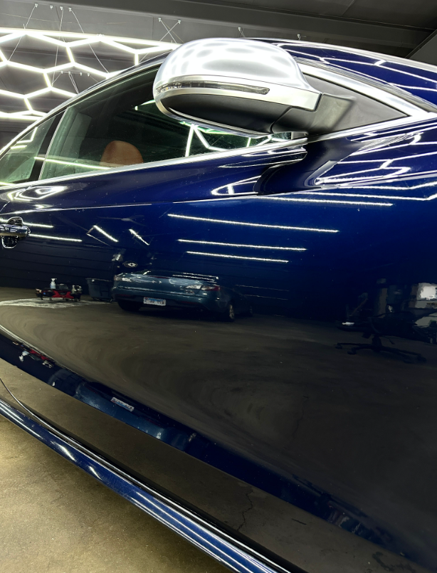 Side view of a glossy dark blue car door reflecting a garage with hexagonal lights, featuring a silver side mirror.