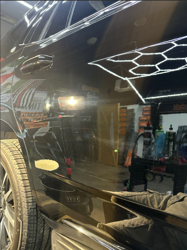 A bright light reflects on the polished black side panel of a car inside a workshop with hexagon ceiling lights.