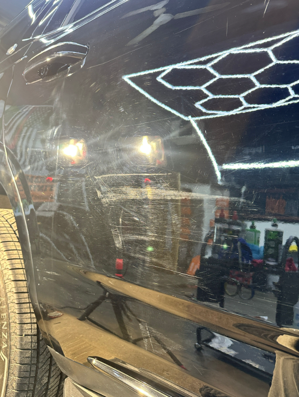 Black car panel with circular swirl marks and surface scratches reflecting shop lights.