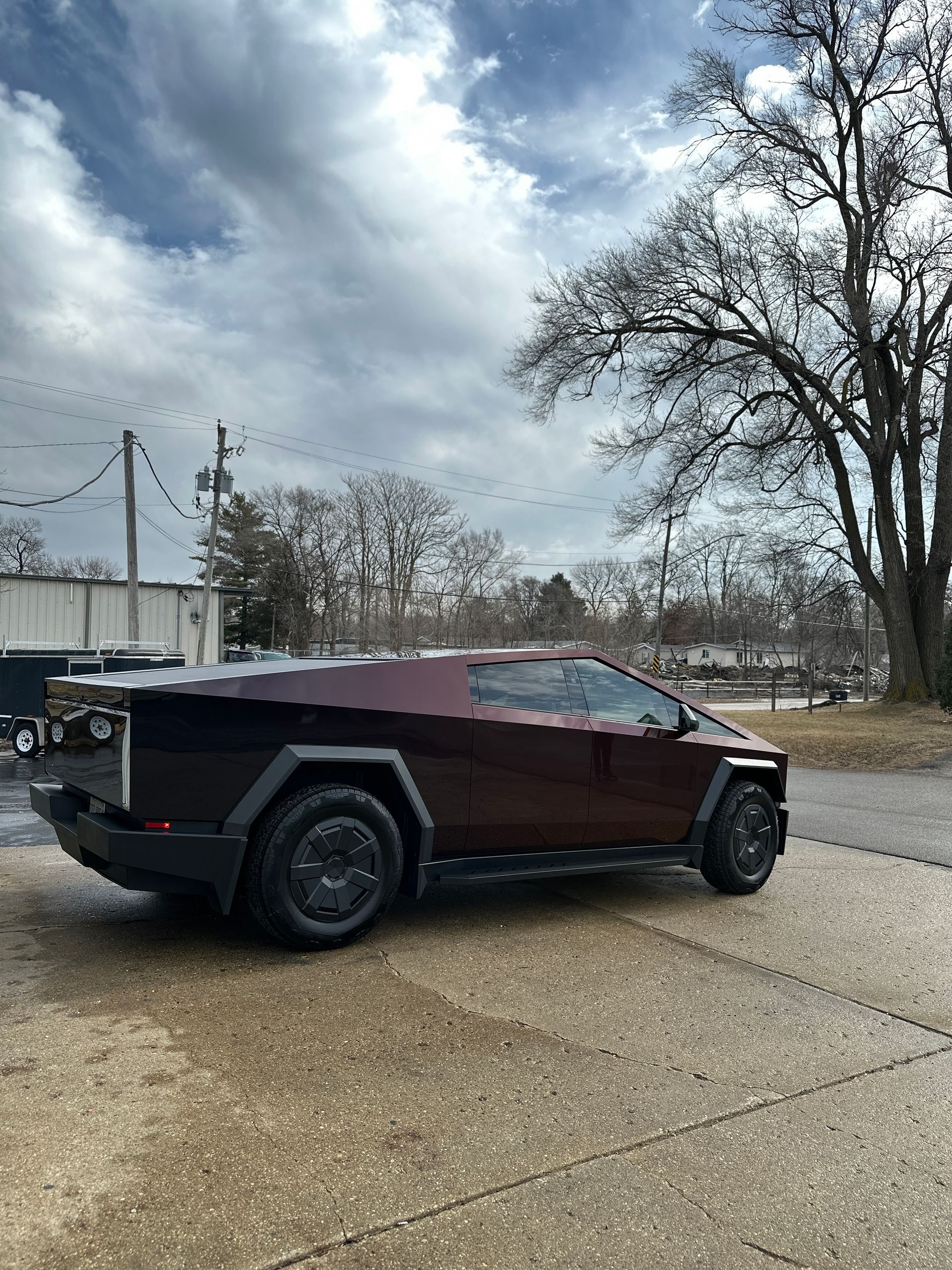 A dark, angular Tesla Cybertruck parked on a gravel lot under a cloudy sky.