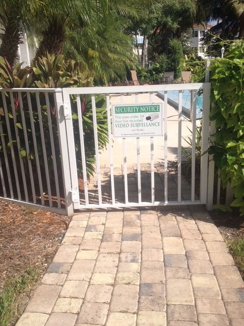 Metal gate at a garden entrance with a sign, bordered by greenery and a stone walkway.
