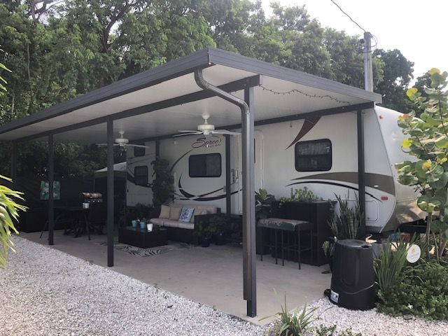 Covered patio beside a parked RV, with outdoor seating, gravel ground, and trees in the background
