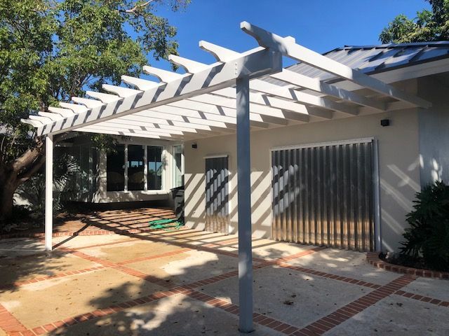 White patio pergola with slatted beams beside a house, casting striped shadows on a brick courtyard