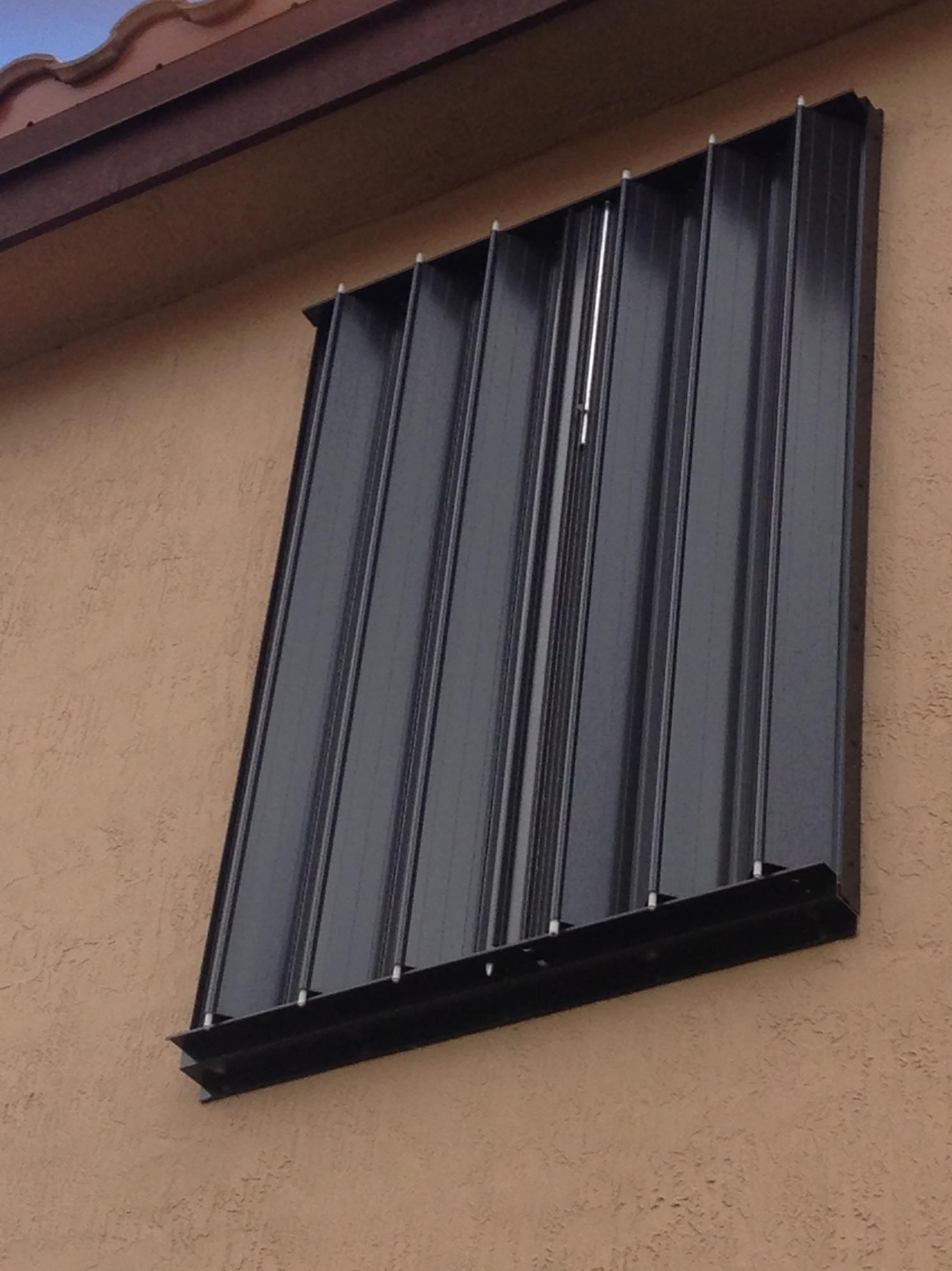 Black metal window shutter mounted on a beige exterior wall under the roof eave