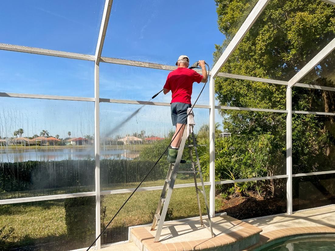 Person standing on a ladder cleaning a screened enclosure beside a lake