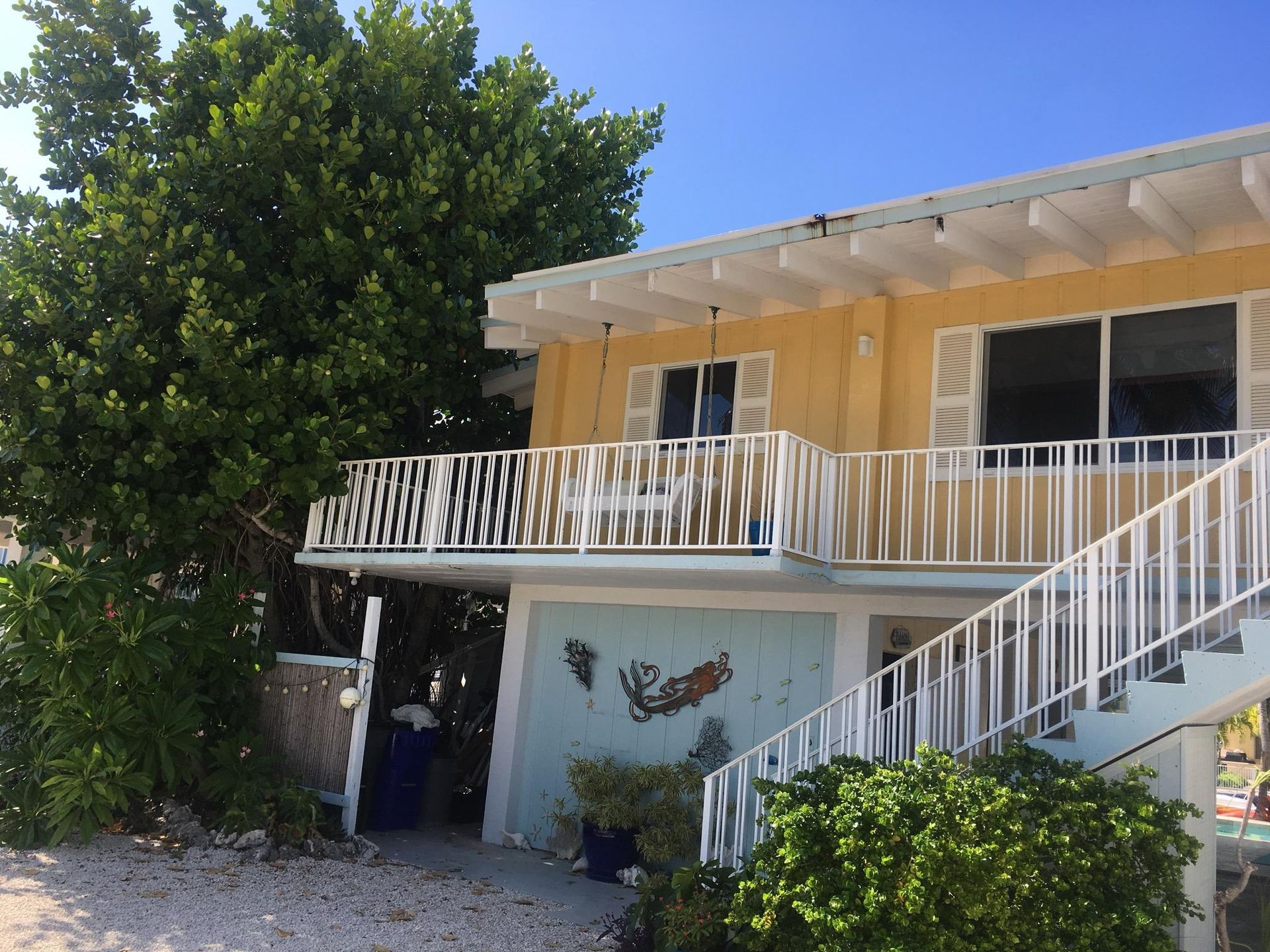 Yellow house with white railings, front porch, and staircase, under a tree on a sunny day