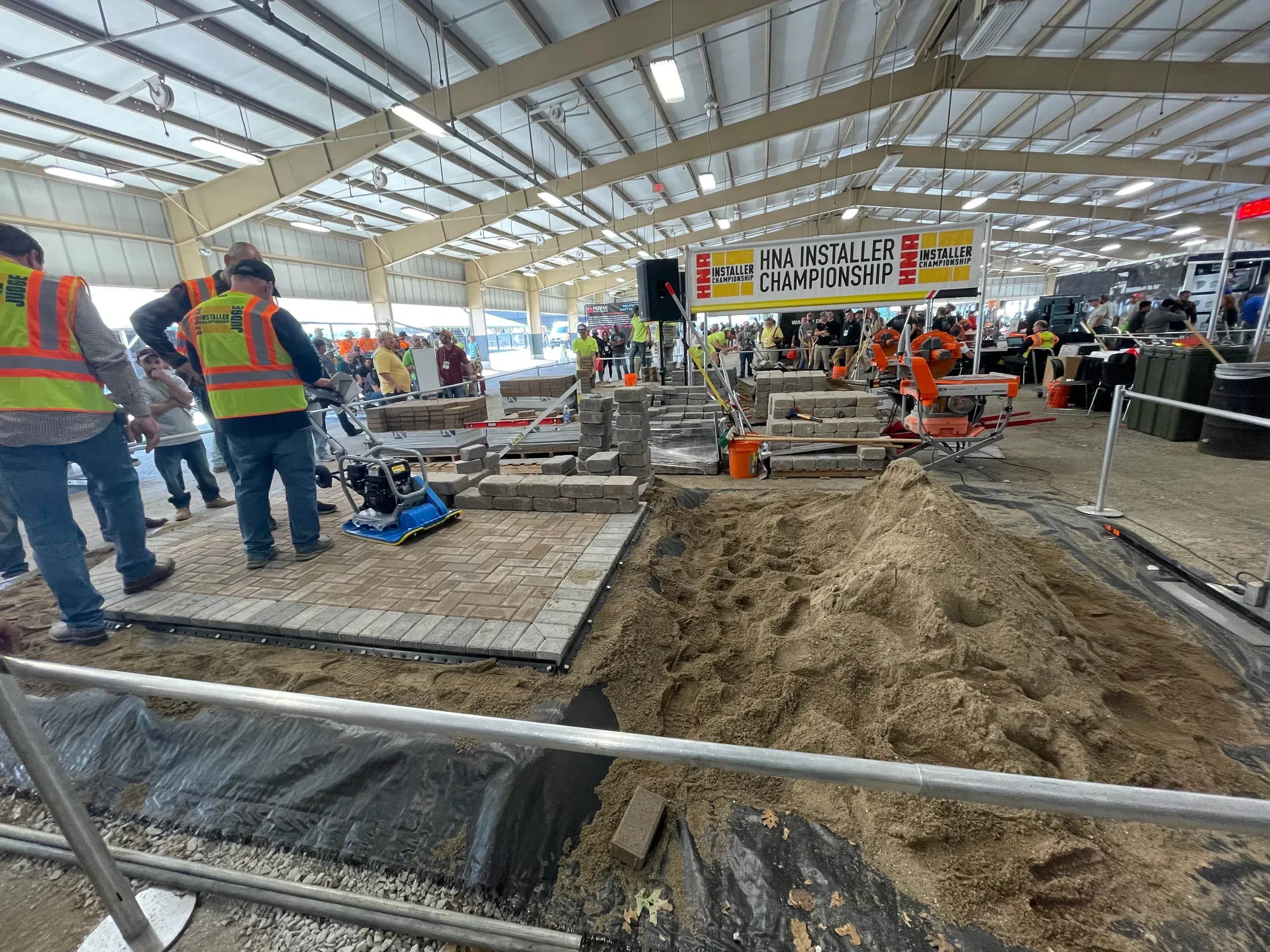 Construction competition: workers laying bricks, large pile of sand, inside an event hall, bright colors, spectators.