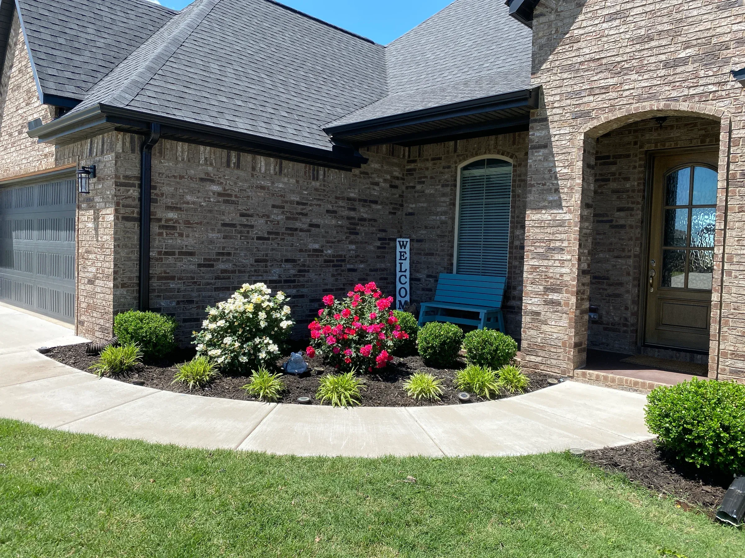 A brick home with a landscaped front yard.  A porch has a bench, surrounded by shrubs, flowers, and a sign.