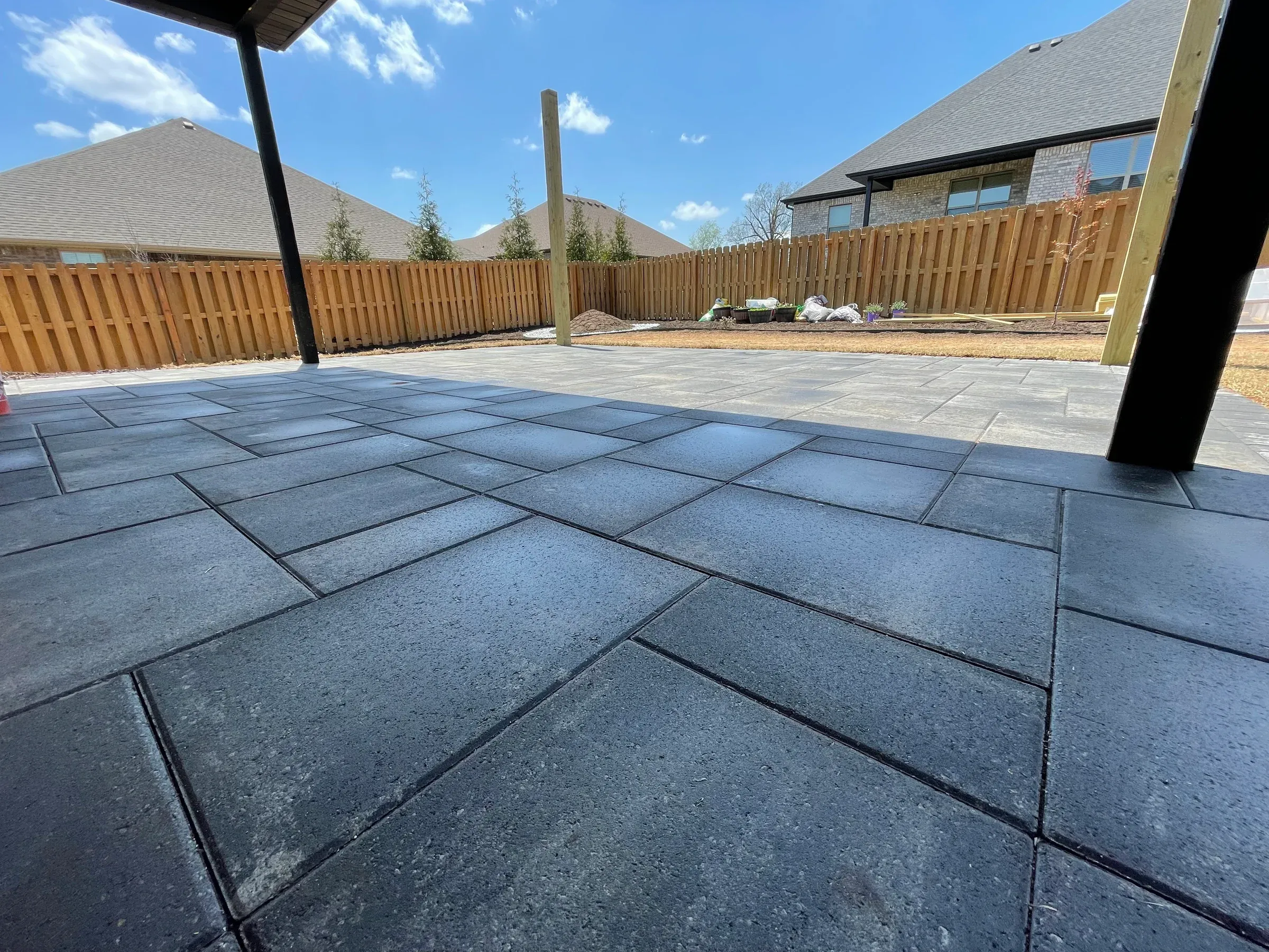 Black pavers on a patio with a wooden fence and houses in the background under a bright blue sky.
