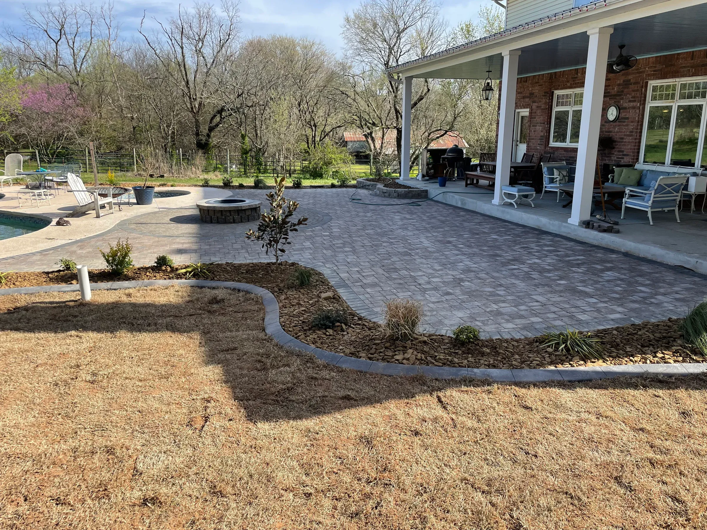 Patio with brick pavers, bordering a lawn with landscaping near a house and pool.