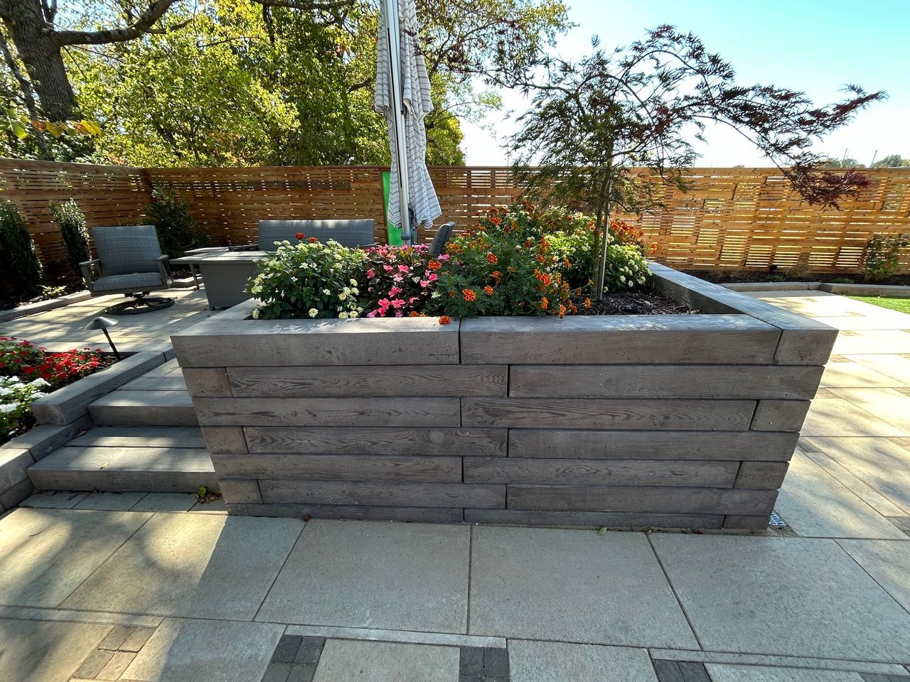 Gray brick planter box filled with flowers sits on a patio. Wooden fence and trees in background.