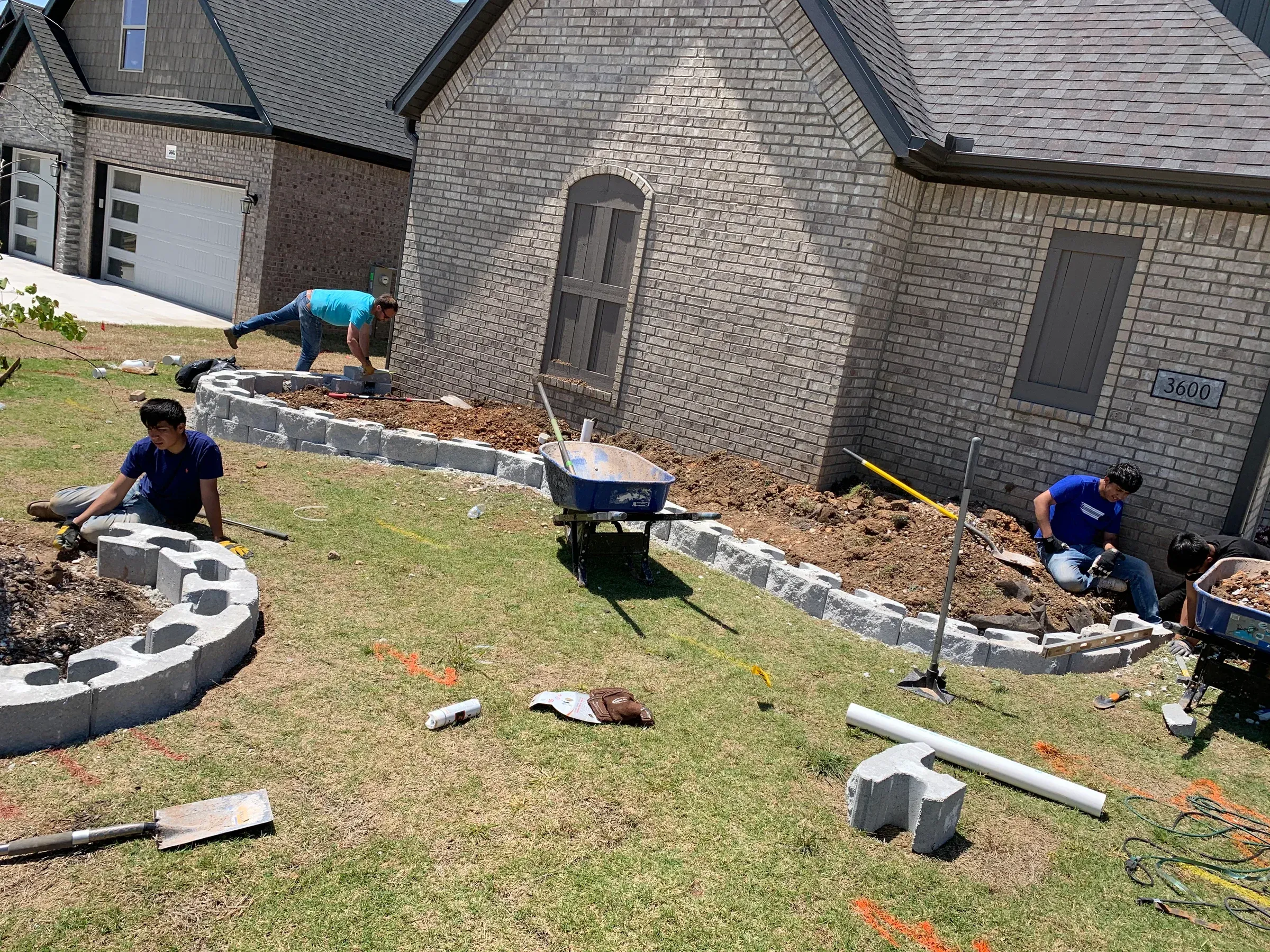 Workers installing landscaping blocks around a house with a brick exterior, on a sunny day.