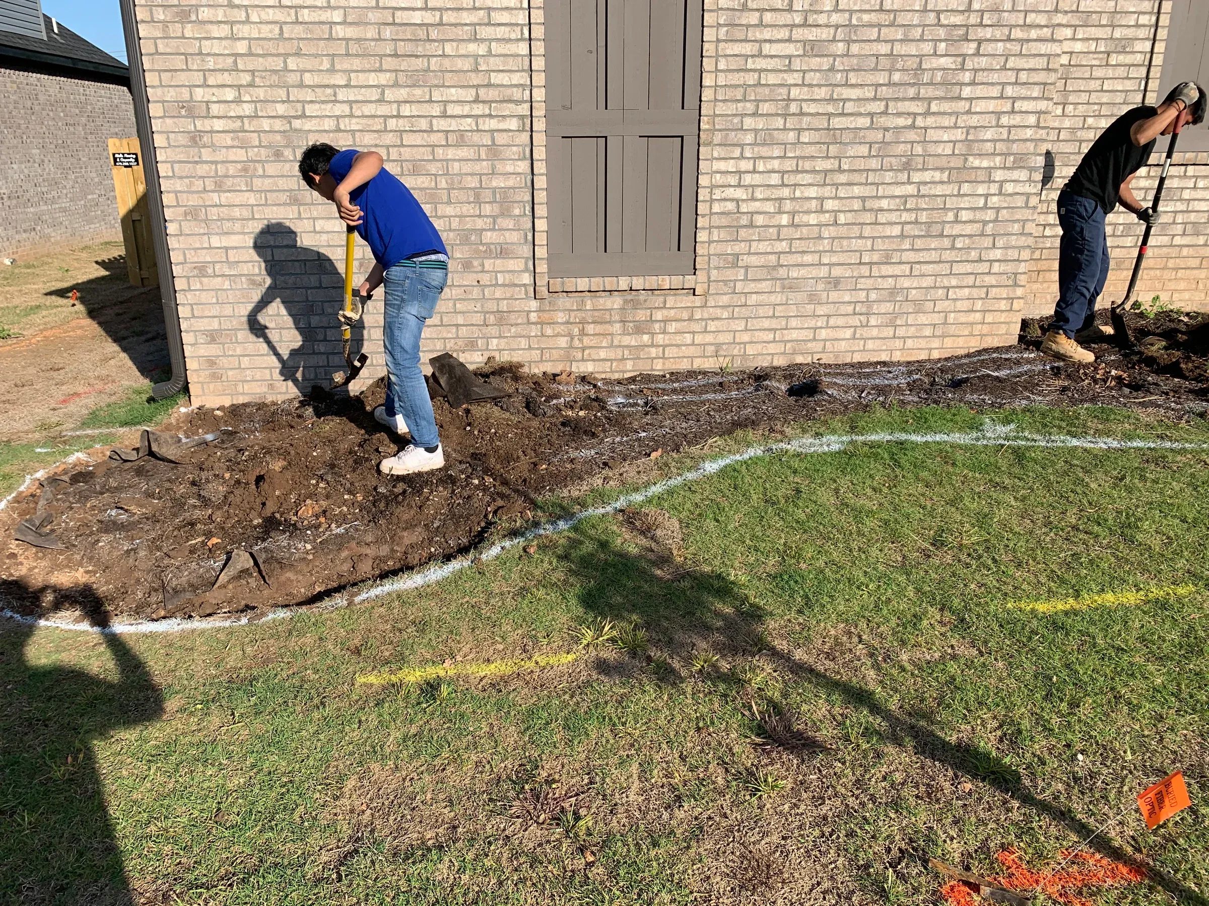 Two people digging in a garden bed near a brick building.