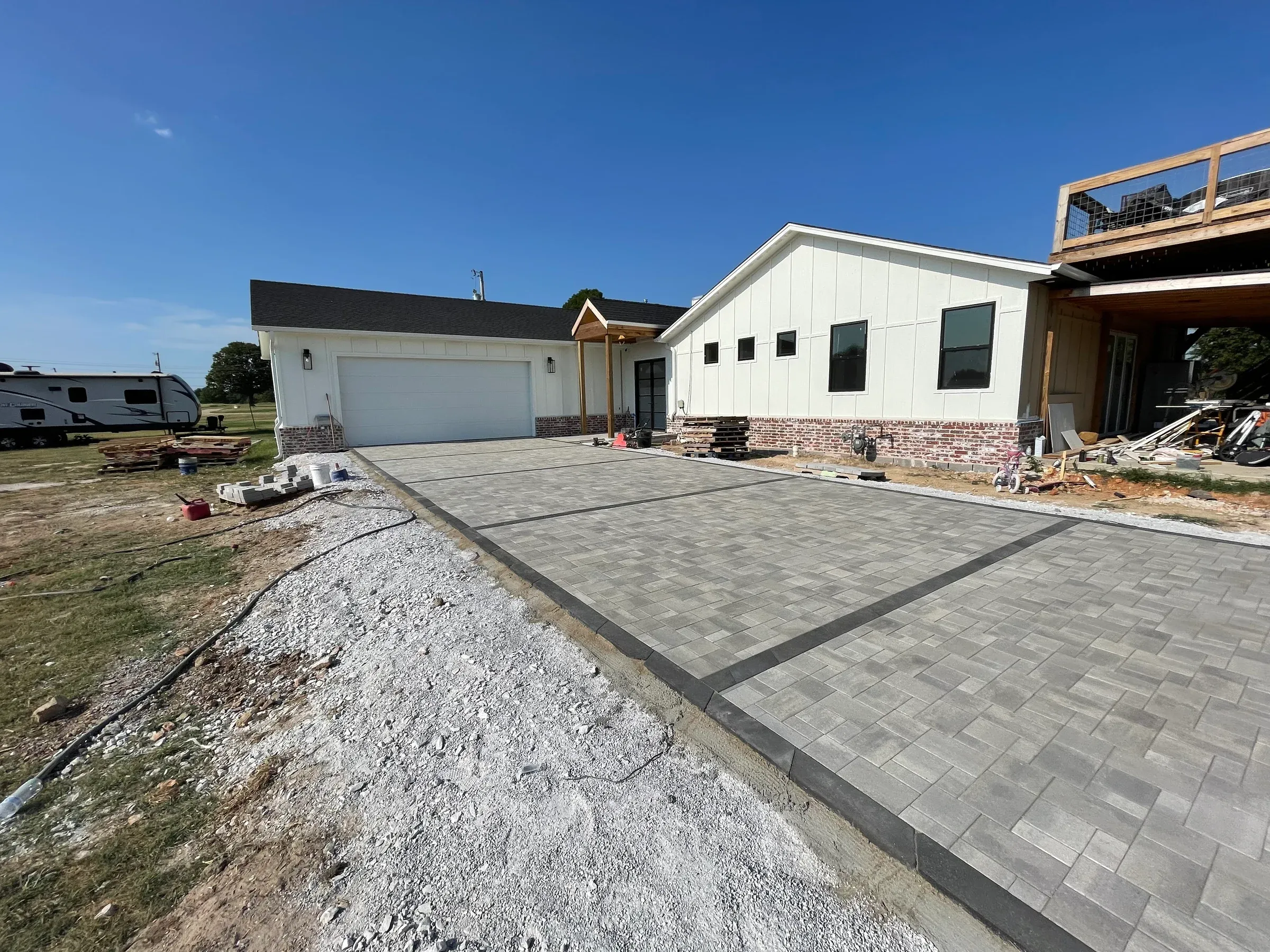 Driveway paving in progress at a white house with a garage and new construction, under a blue sky.