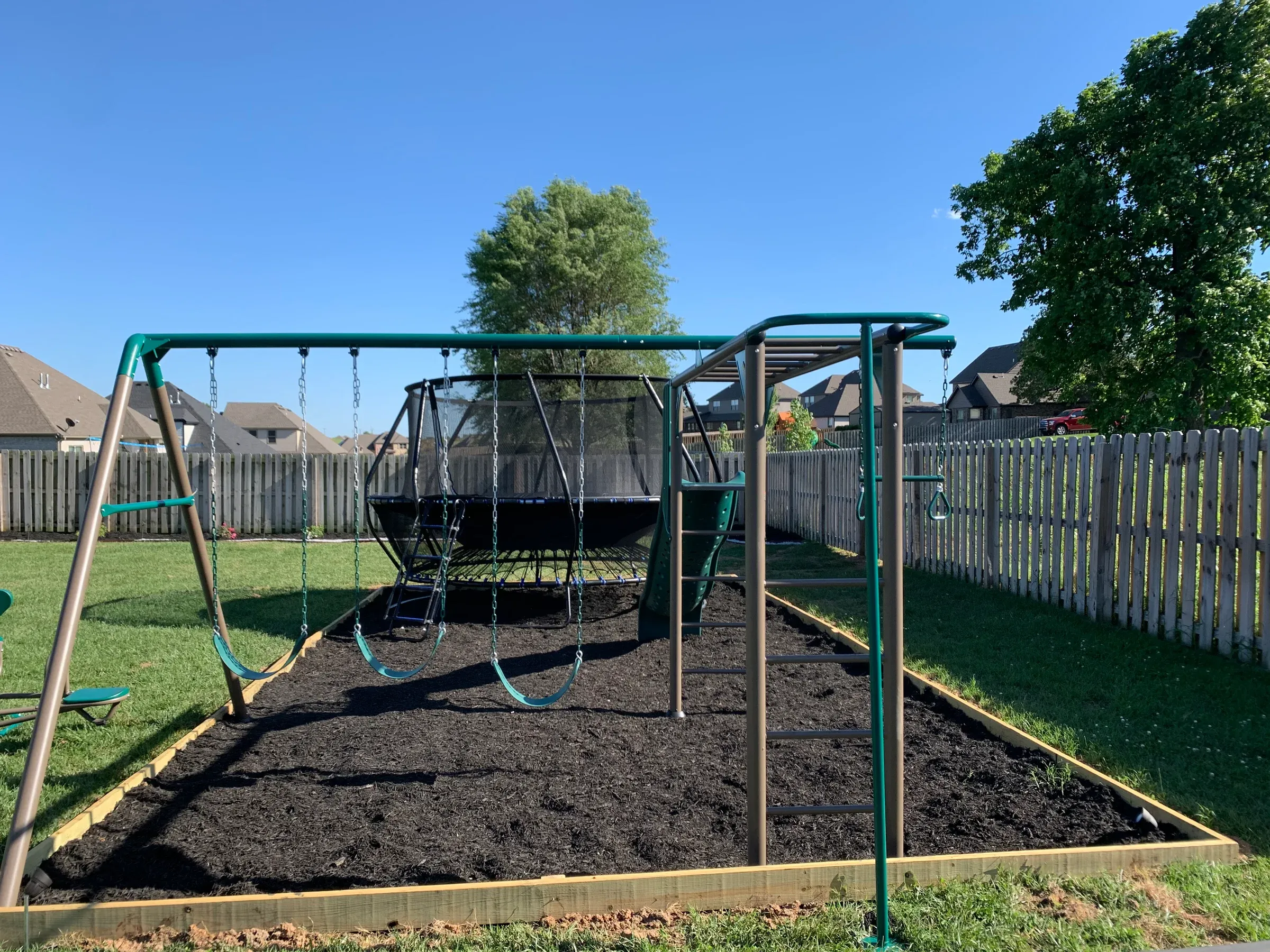 Playground with swings, trampoline, and ladder on a mulched area, bordered by a fence and houses.