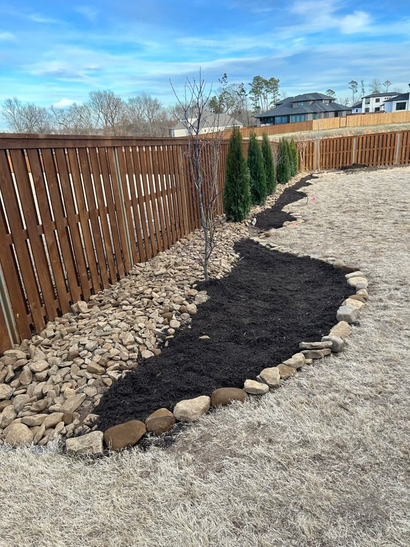 Backyard garden bed with dark mulch, rocks, evergreens, and wooden fence.