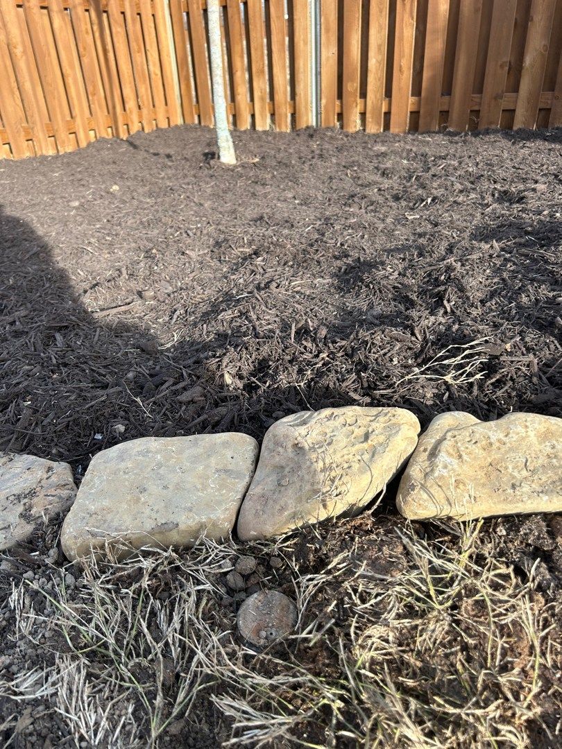 Rocks border a mulched flower bed next to a wooden fence.
