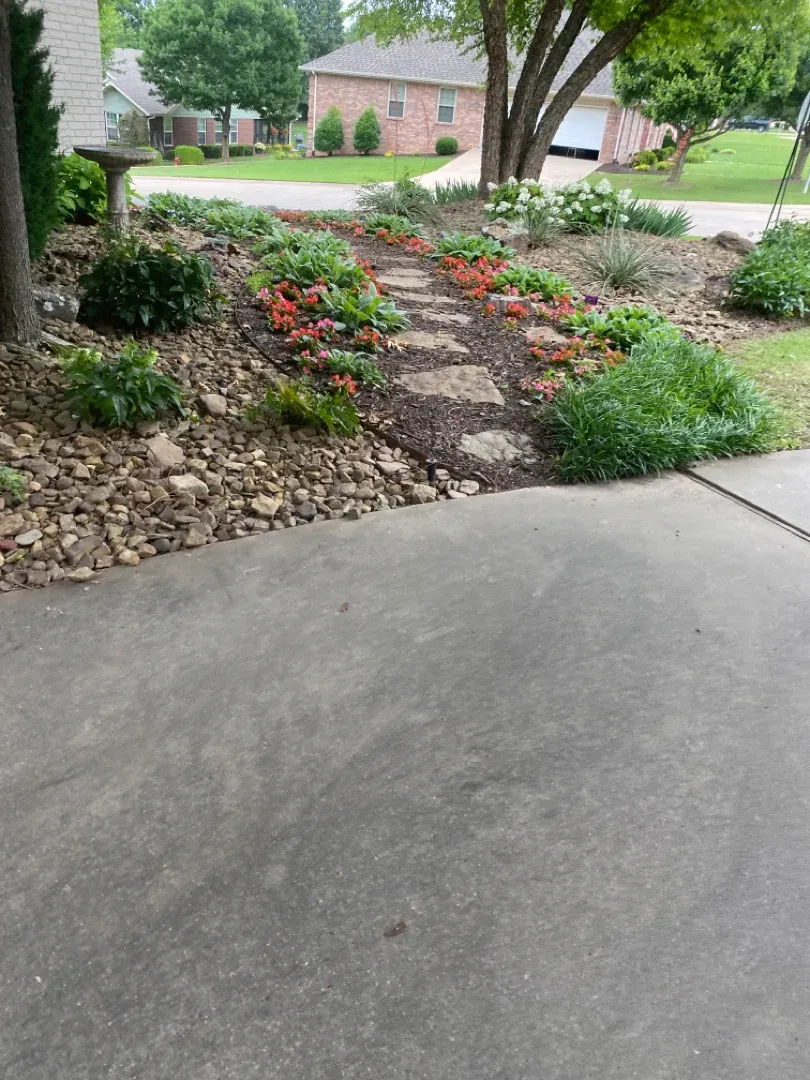 Concrete driveway leads to a garden bed with stone steps, various flowers, and a house in the background.
