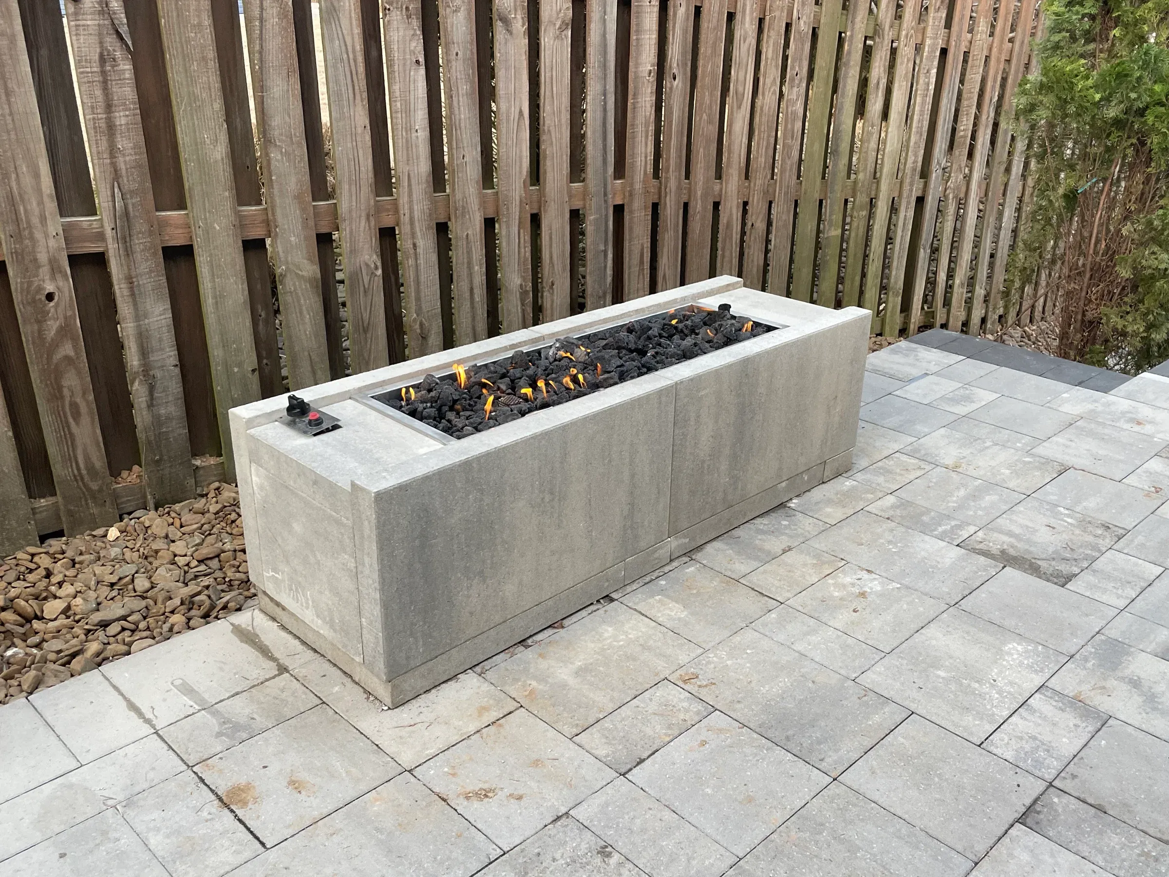 Rectangular gray concrete fire pit with burning flames, on a paved patio near a wooden fence.