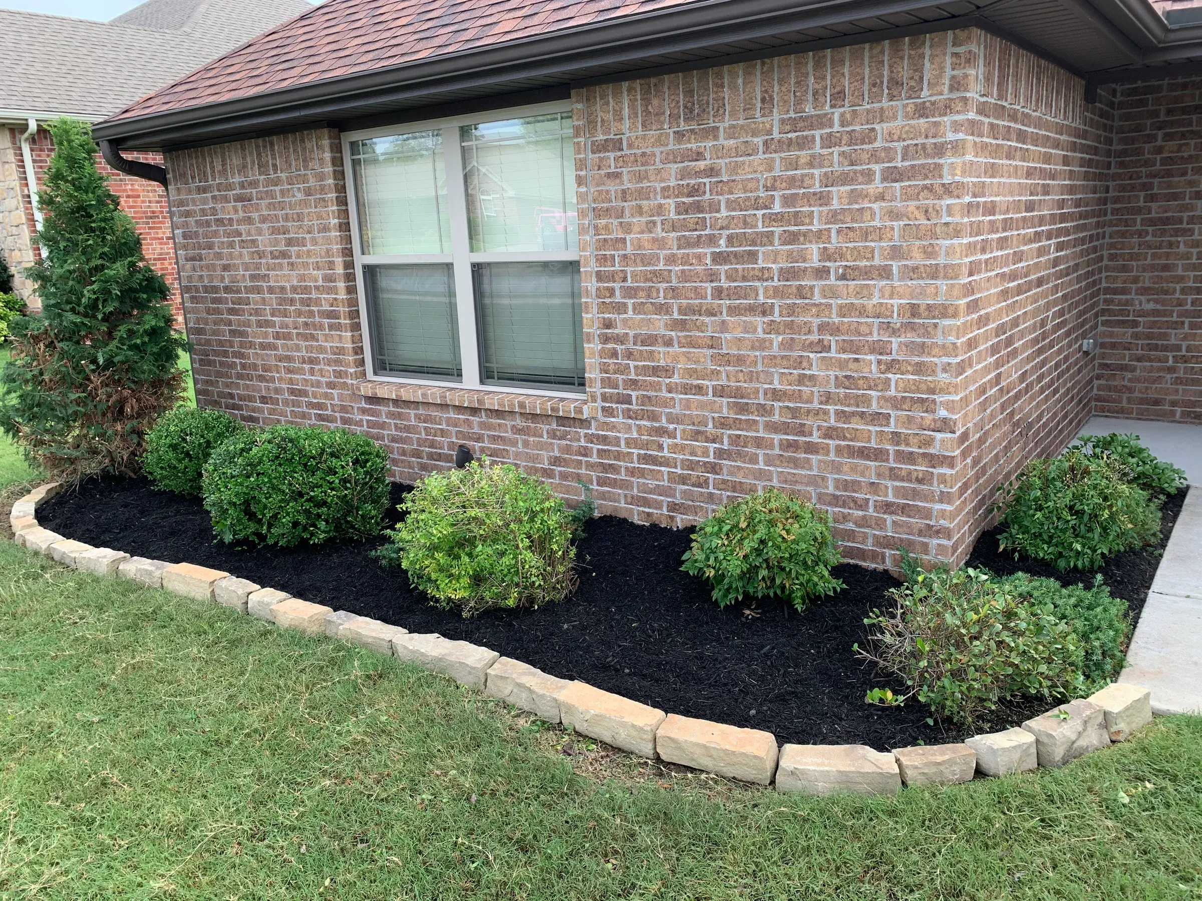 Landscaped yard with brick house, window, green grass, and mulch-filled flower bed bordered by stone.