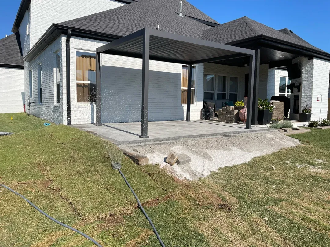 Backyard patio with pergola, brick house, and green grass. Grey stone patio and grey pergola frame.