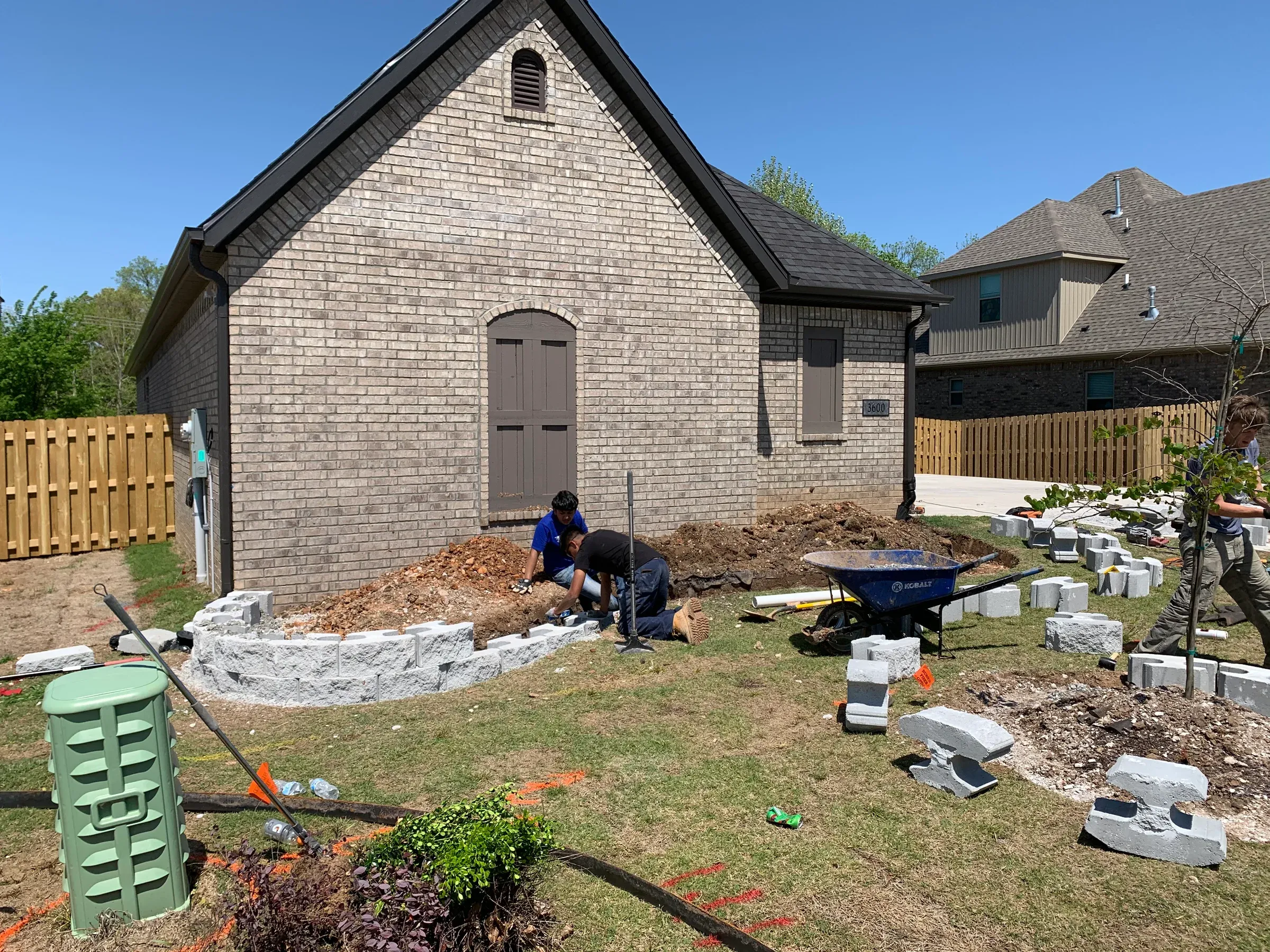 Two people working on landscaping near a brick building. Construction materials and a wheelbarrow are present.