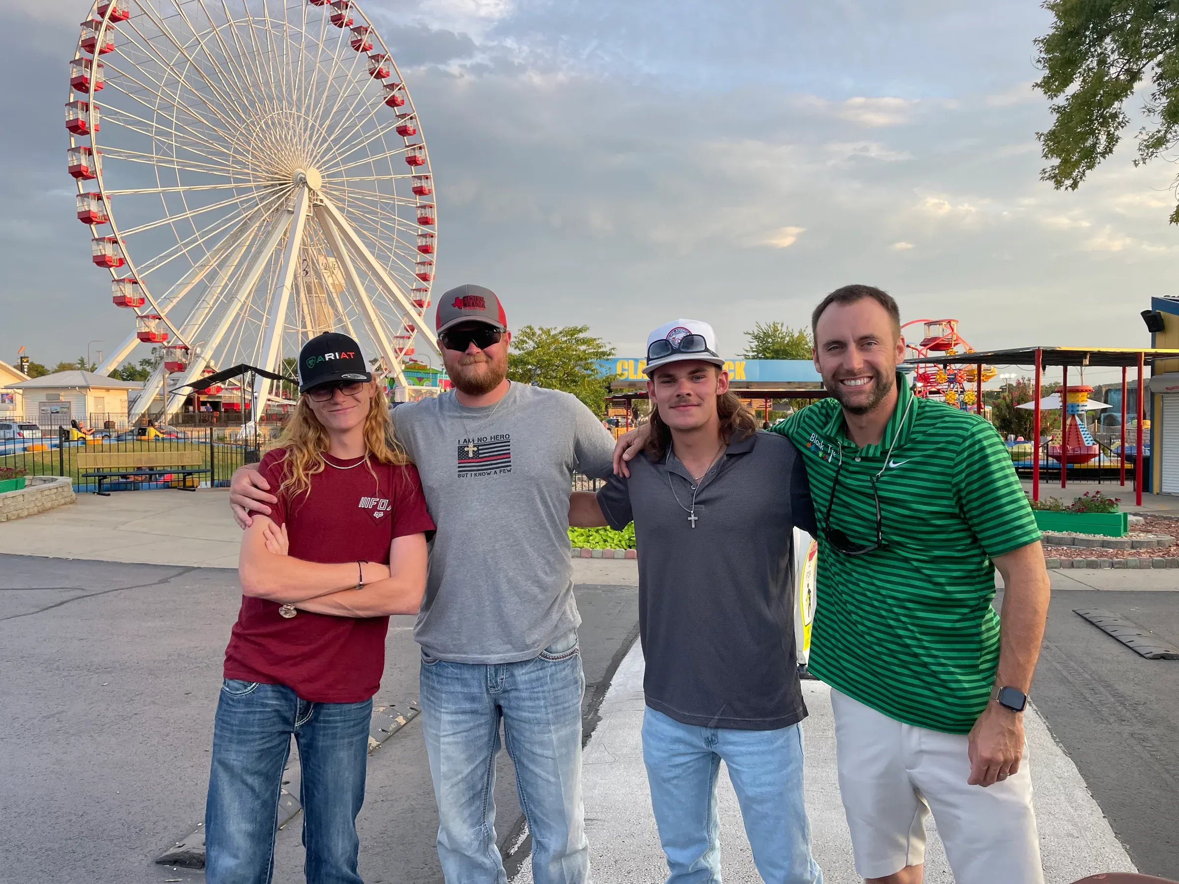 Four men pose in front of a Ferris wheel. One in red shirt with arms crossed, others in casual wear. Cloudy sky.