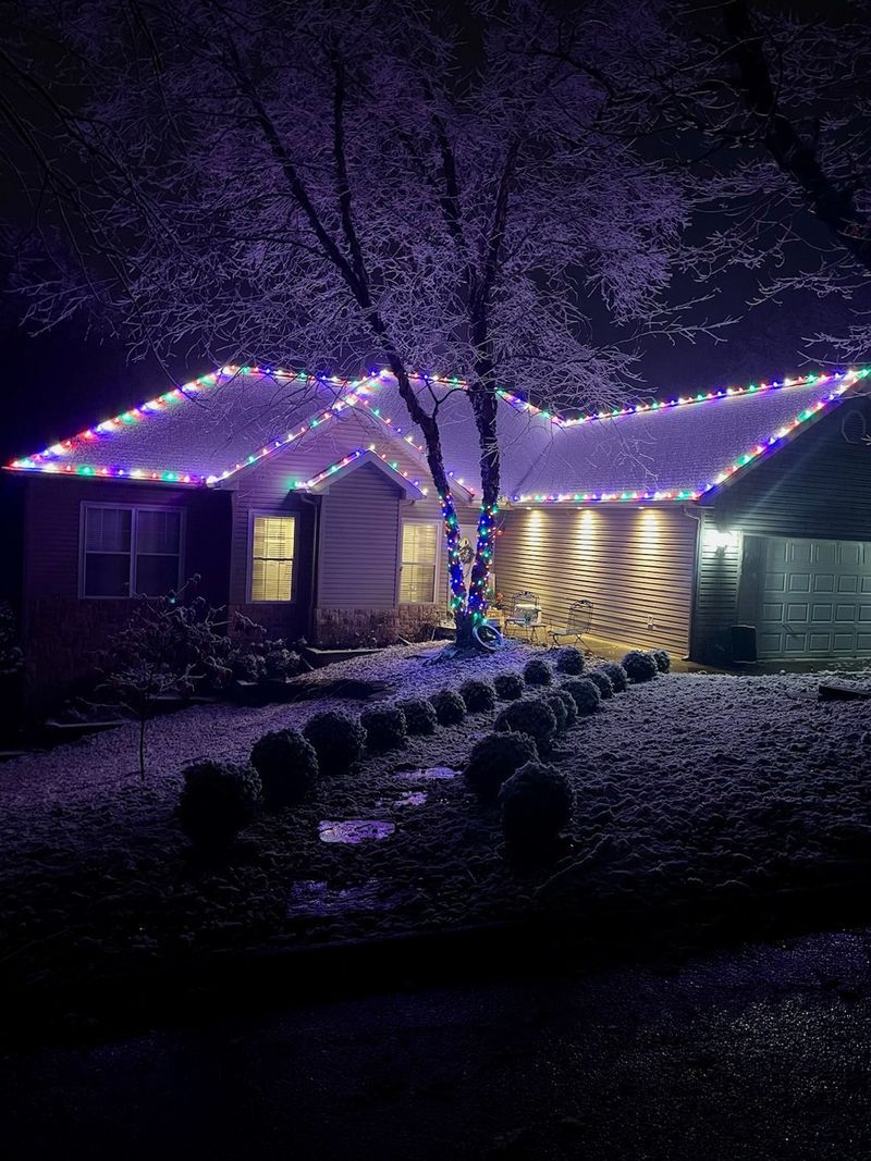 House at night with roof and tree wrapped in purple and white Christmas lights.