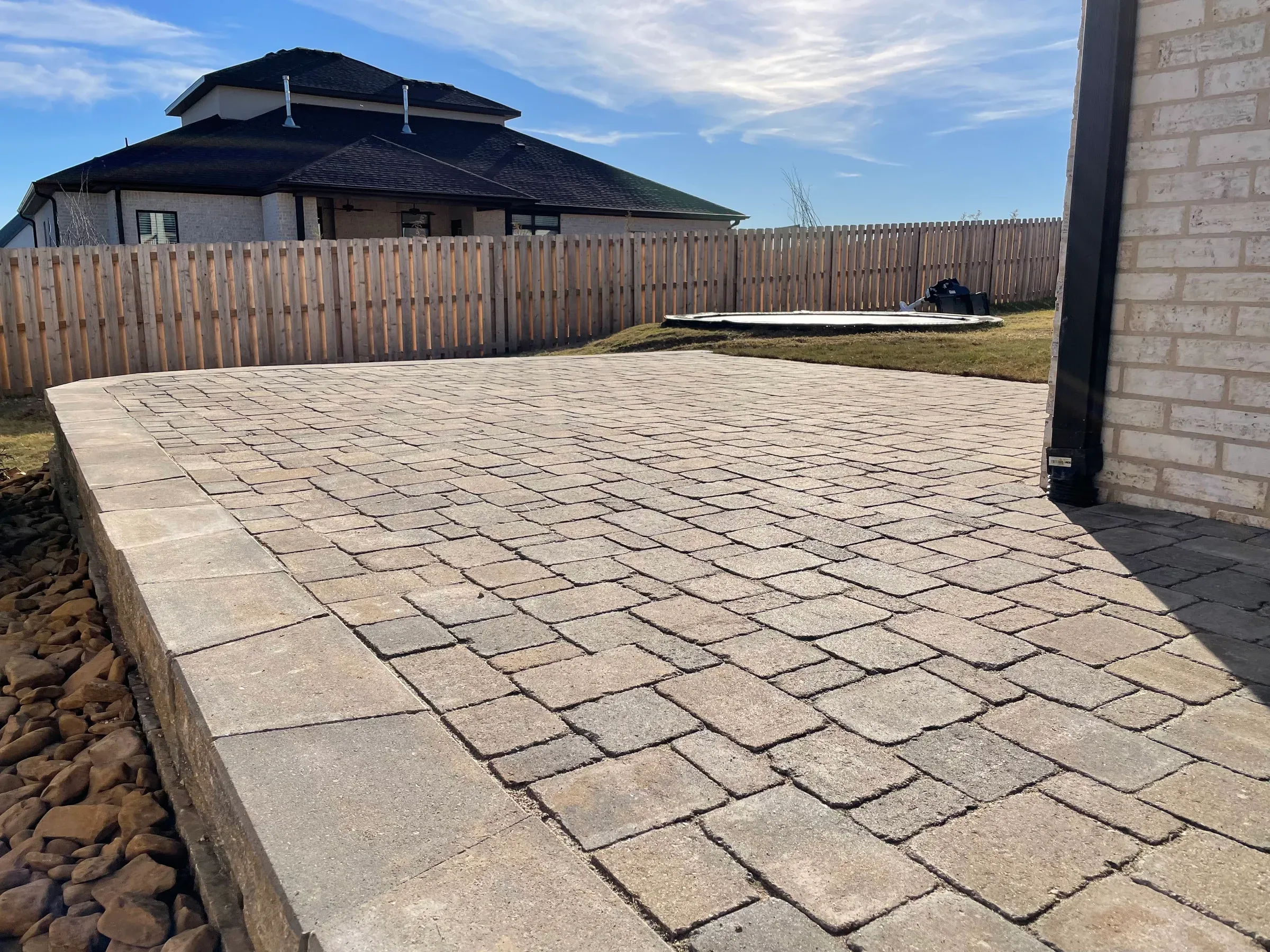Stone patio and retaining wall in a backyard with a wooden fence and house in the background.