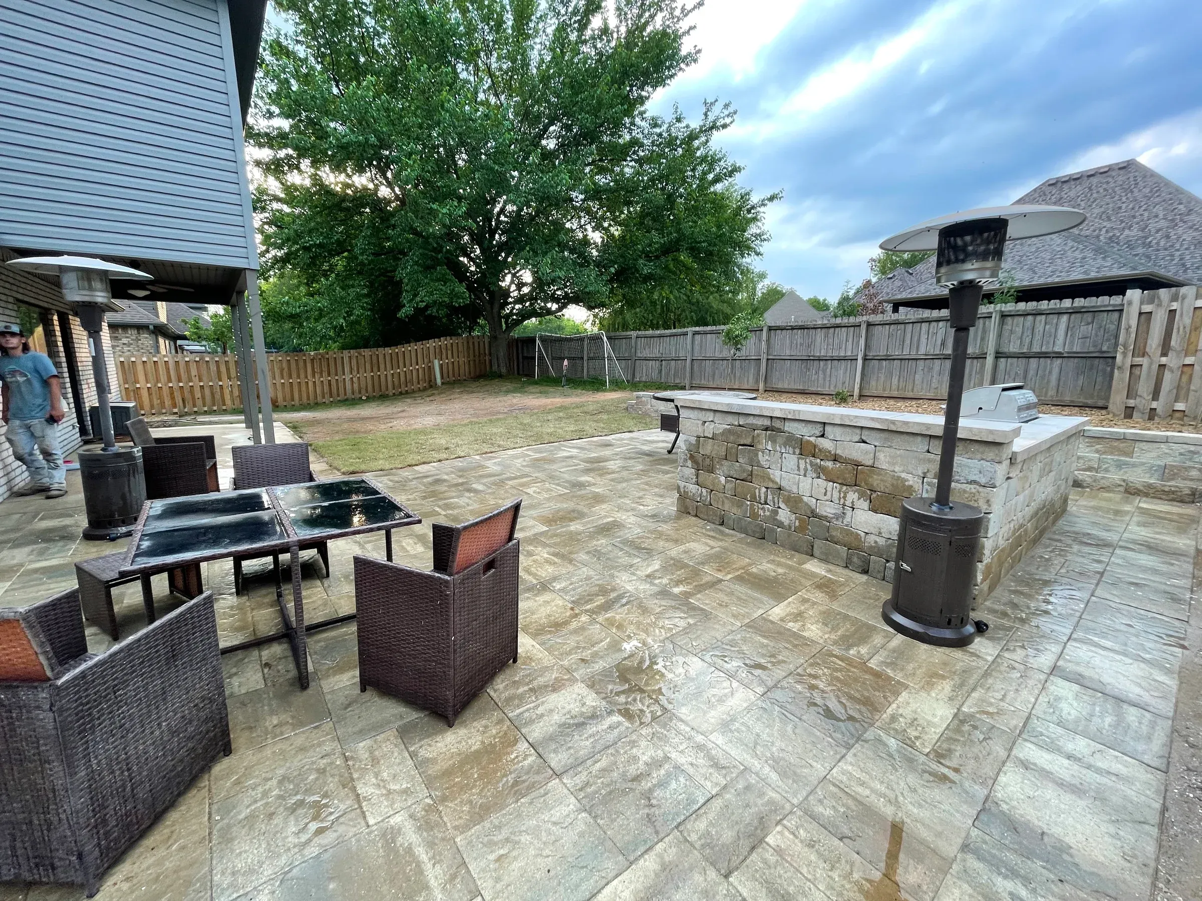 Patio with stone pavers, outdoor kitchen, seating area, and tree-lined yard under a cloudy sky.