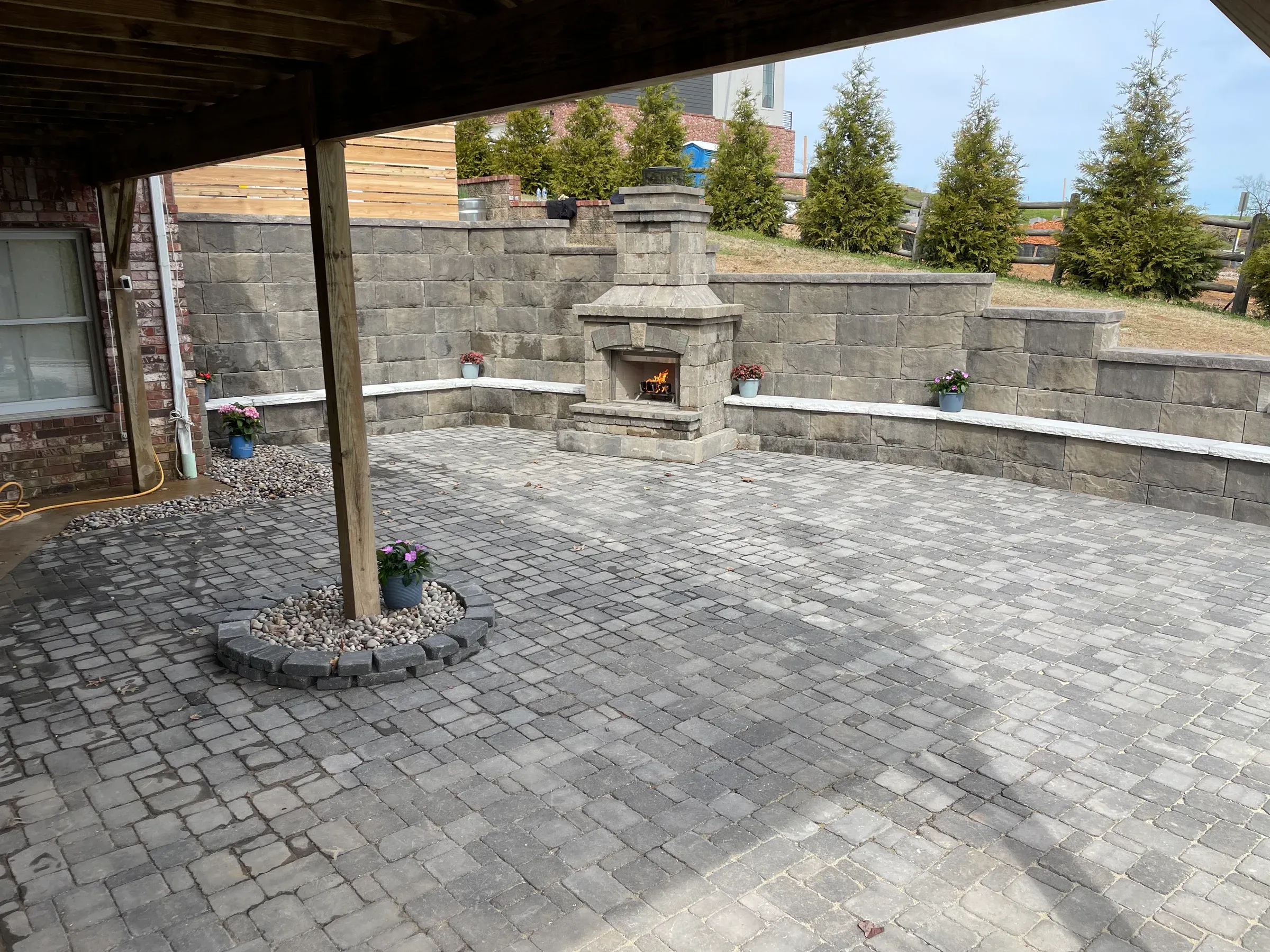 Patio with stone pavers, fireplace, and retaining wall. Underneath a wooden structure, with trees in the background.