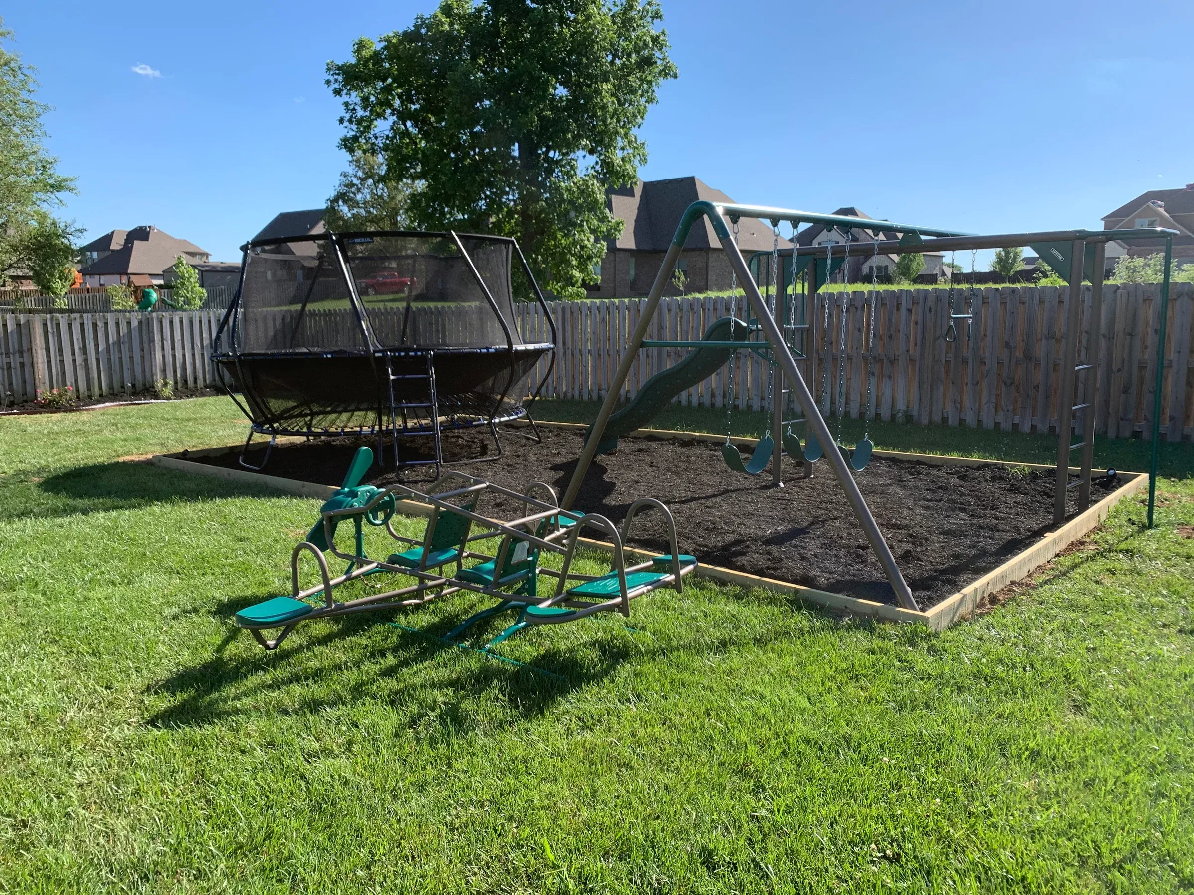 Backyard playground with a swing set, trampoline, and seesaw on a bed of dark mulch.