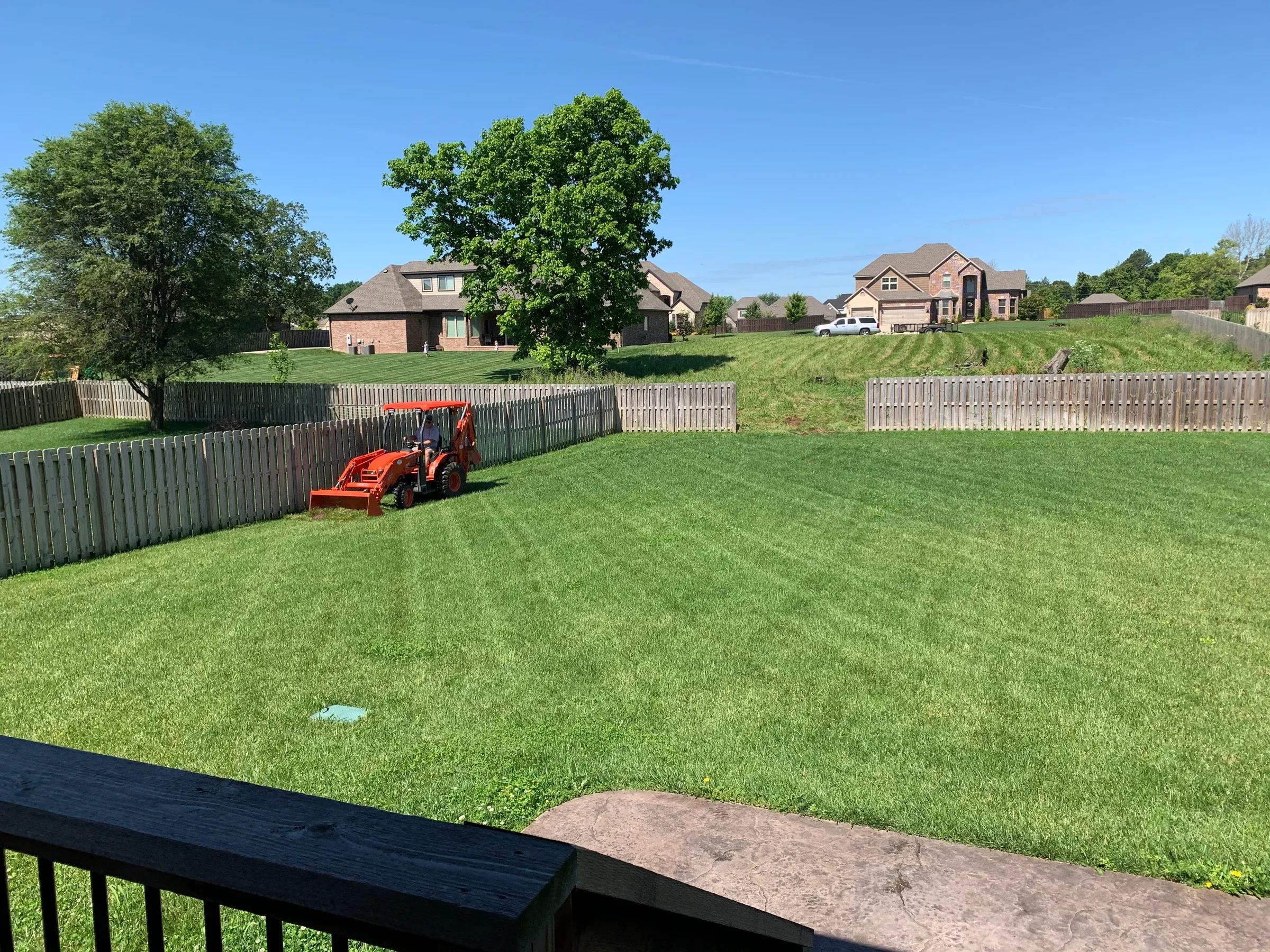 Orange tractor mowing a green lawn in a fenced backyard on a sunny day. Houses in the background.