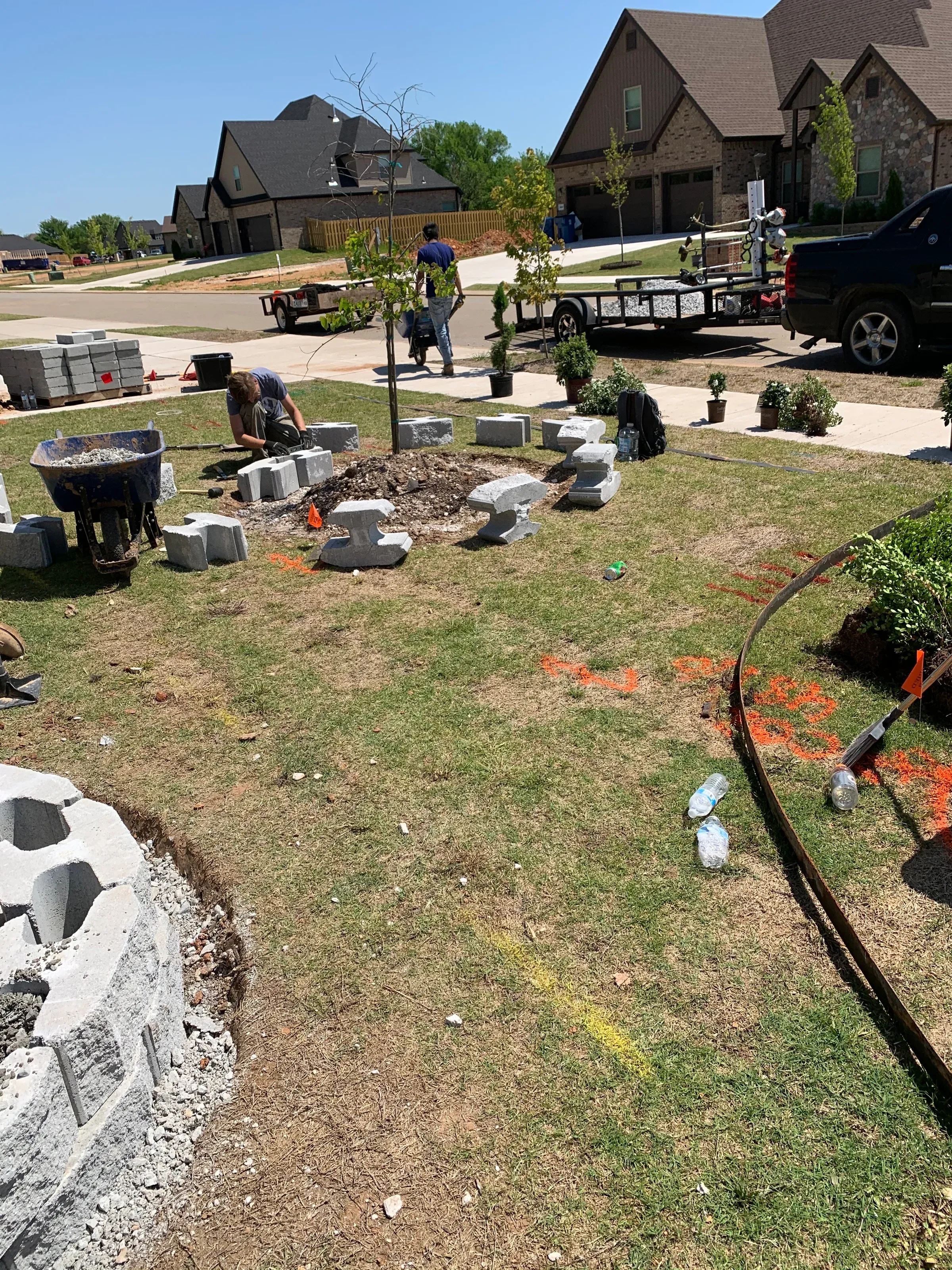 Workers arranging stone blocks around a small tree in a grassy yard, surrounded by tools and materials.