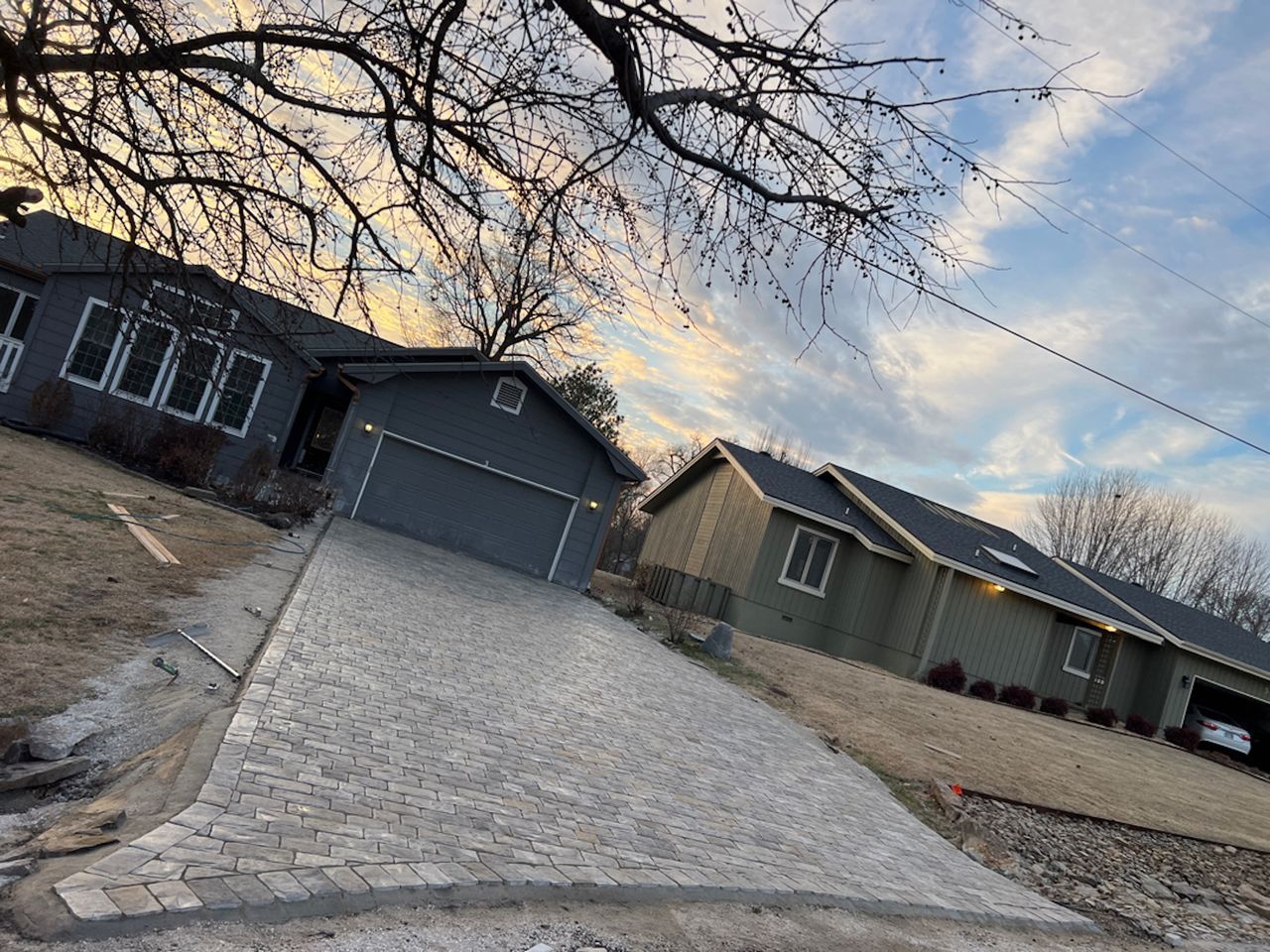 Driveway slopes toward two houses under a blue sky. One house is gray, the other green. Bare tree branches overhead.