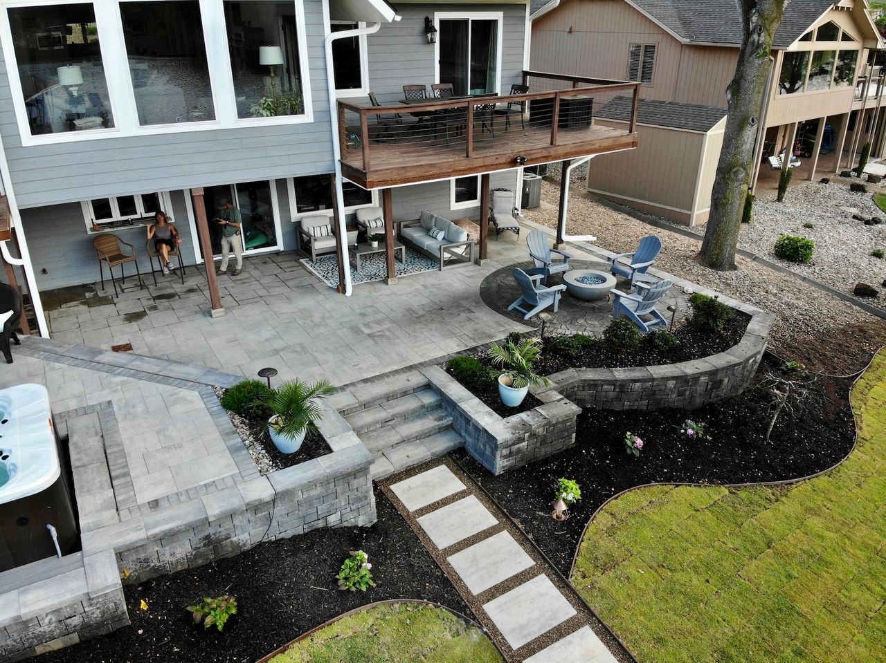 Patio with retaining walls, fire pit, deck, and lake house. Gray and brown tones, outdoor setting.