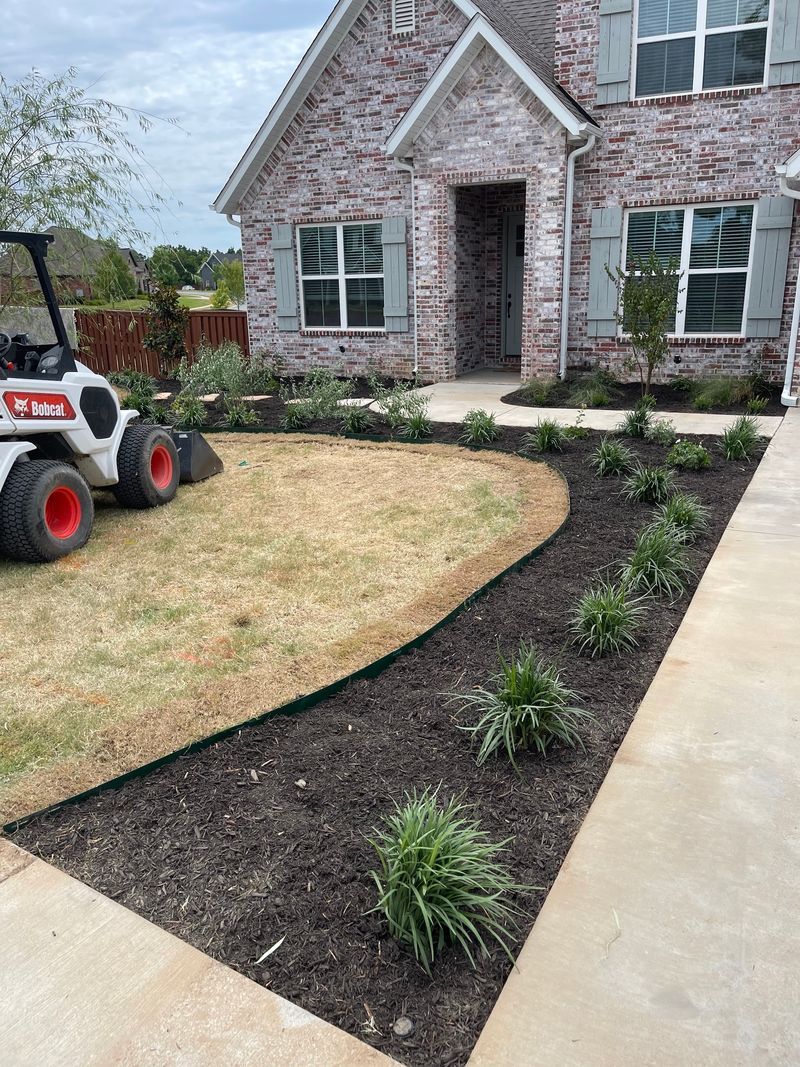 A house with brick facade and landscaping, featuring a lawn, flowerbed, and small shrubs. A Bobcat tractor is in the yard.