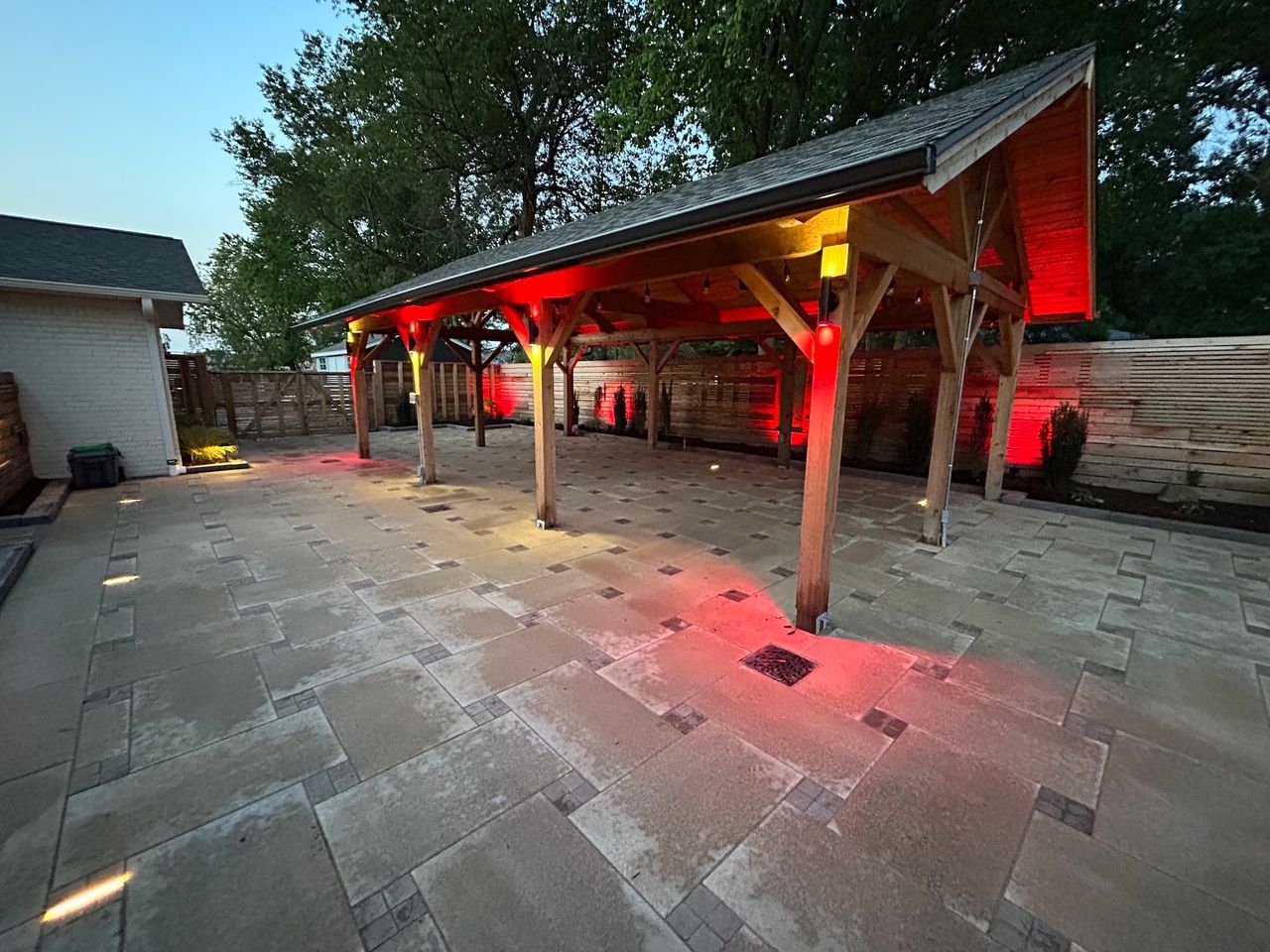 Patio with a wooden pergola lit with red and yellow lights. Stone pavers on the ground.