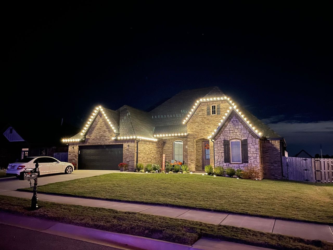 House with white Christmas lights on roofline, at night. White car parked in the driveway.