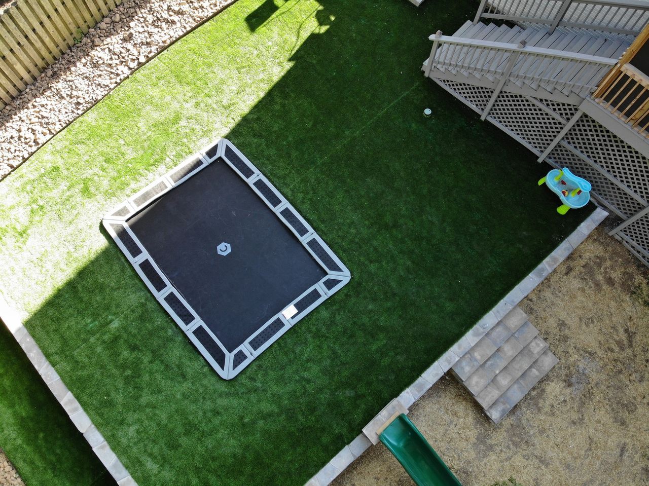 Overhead view of a rectangular trampoline set into a green lawn, next to a deck and small slide.