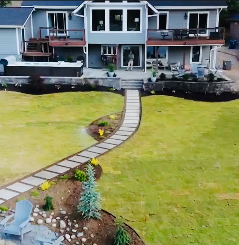Large gray house with a pathway through a green lawn. Hot tub and deck. Person standing by the door.